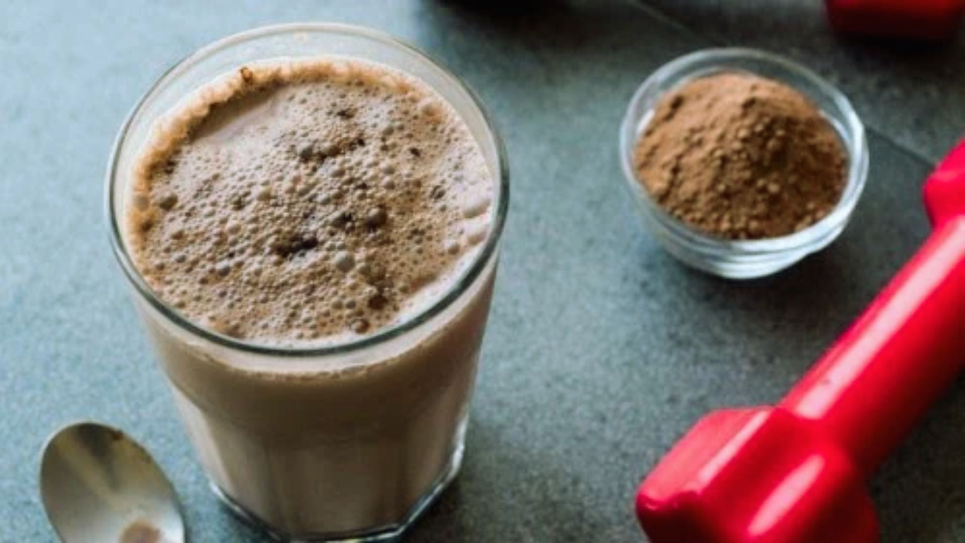 A glass of chocolate shake beside a spoon and two dumbbells on a table, suggesting a balance of indulgence and fitness.