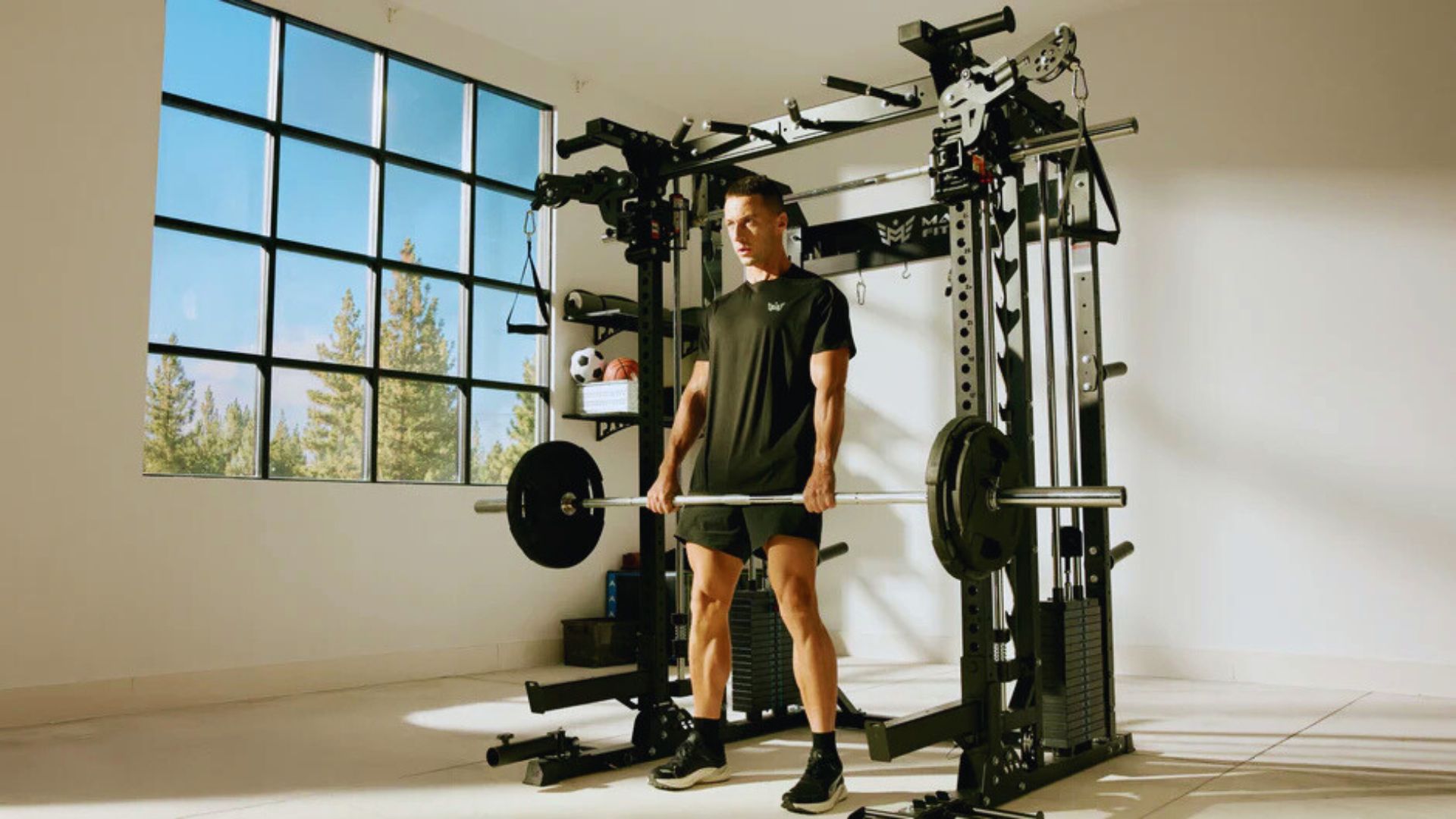 A man in athletic wear performs a barbell deadlift in a sunlit home gym. The room has a large window with a view of trees, creating a calm yet focused atmosphere.