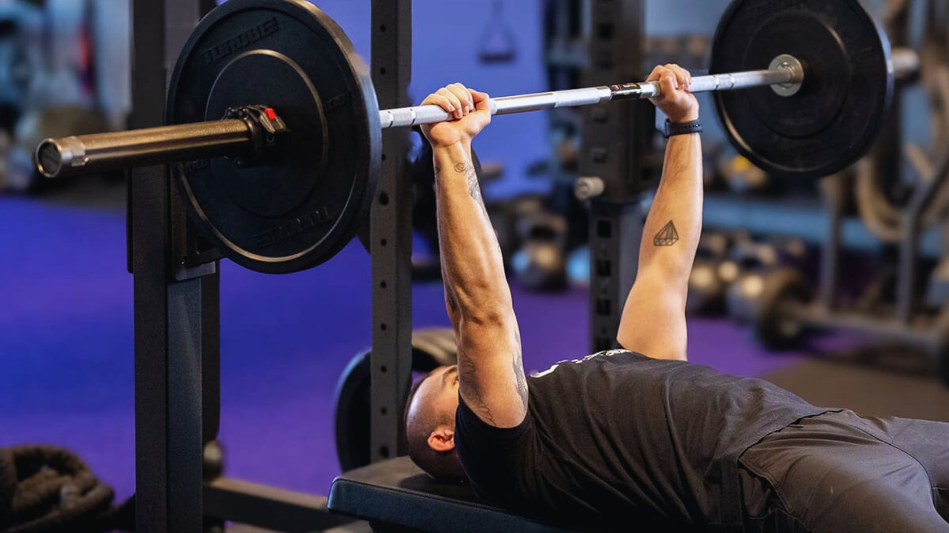 A man bench presses a barbell in a gym, focusing intensely. The gym has a blue-purple hue, with weights and equipment visible in the background.