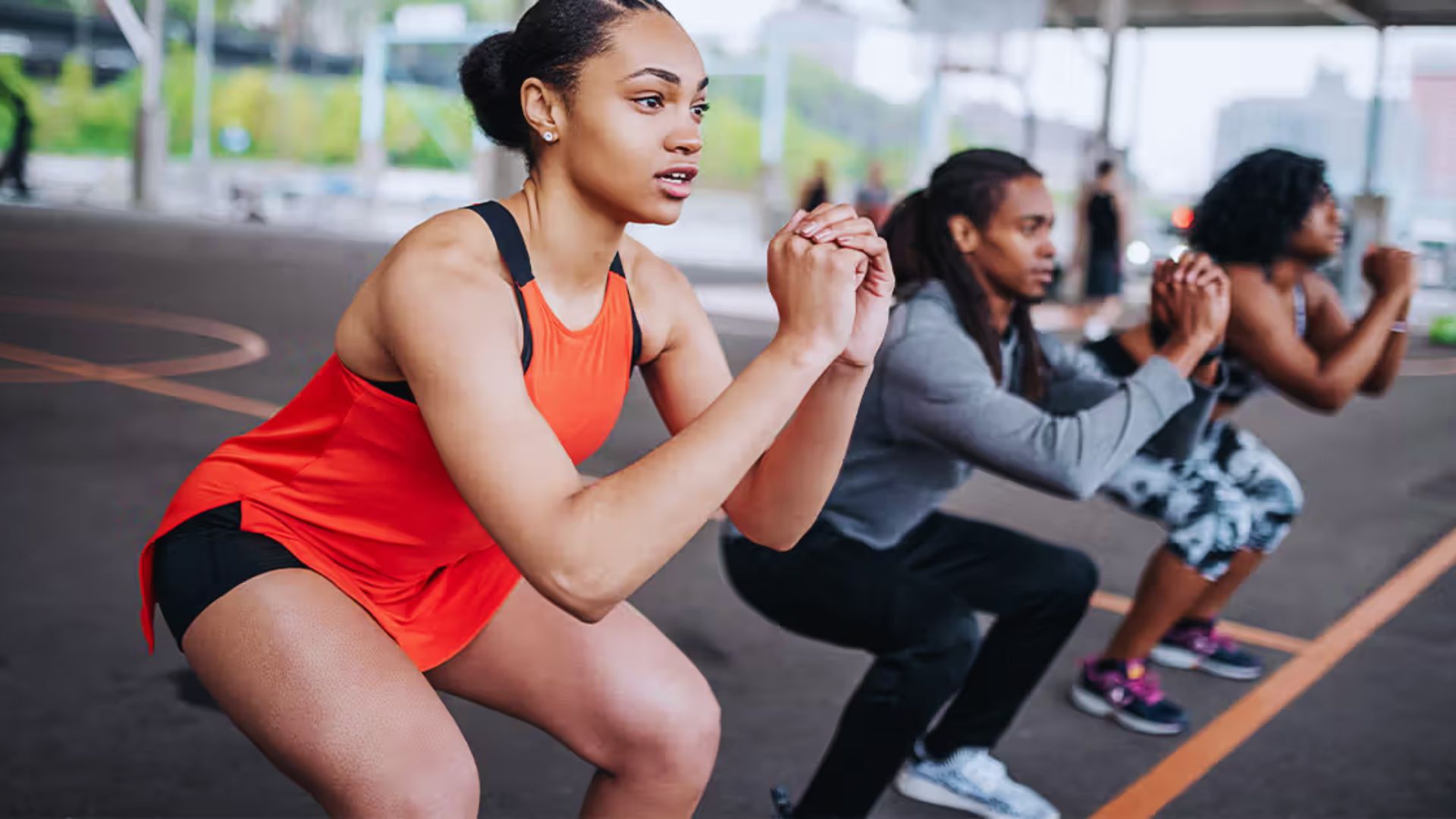 A group of three people exercises outdoors, performing squats. The focus is on a woman in a red tank top, showcasing determination and teamwork.