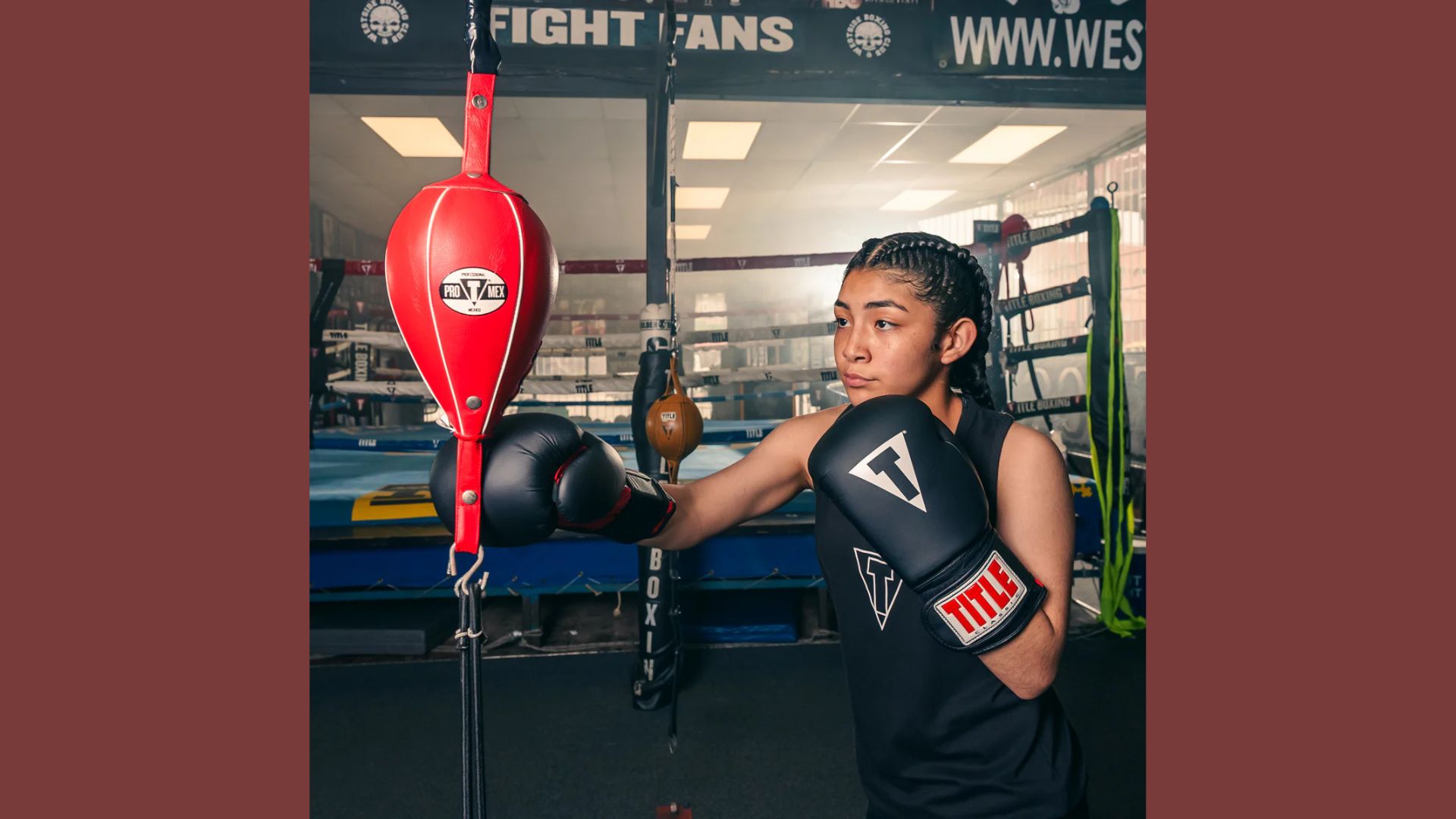A young woman in boxing gear stands confidently, holding a punching bag in a training environment.