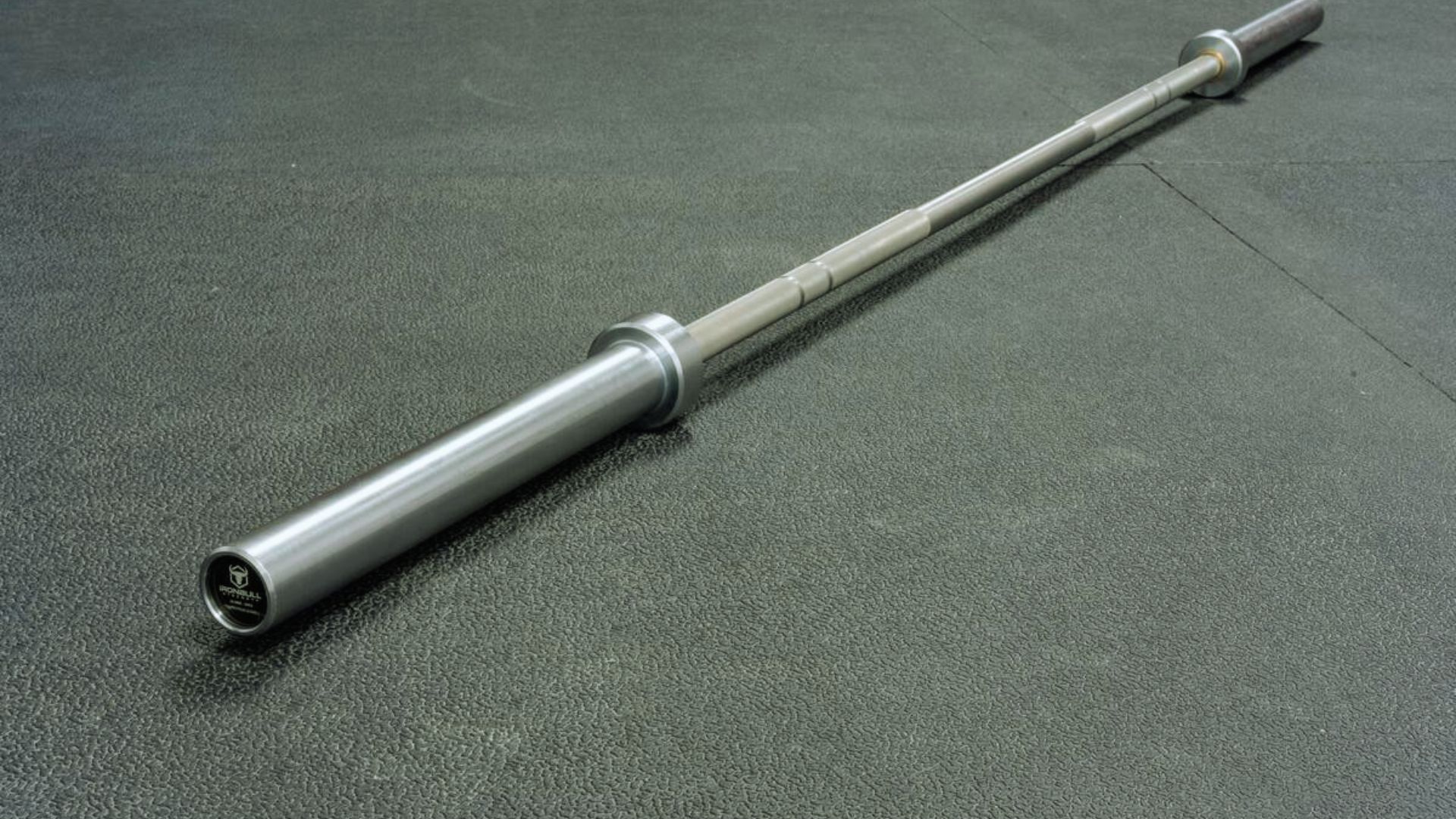 A barbell rests on the floor next to a metal rod, indicating a weightlifting setup in a gym environment.