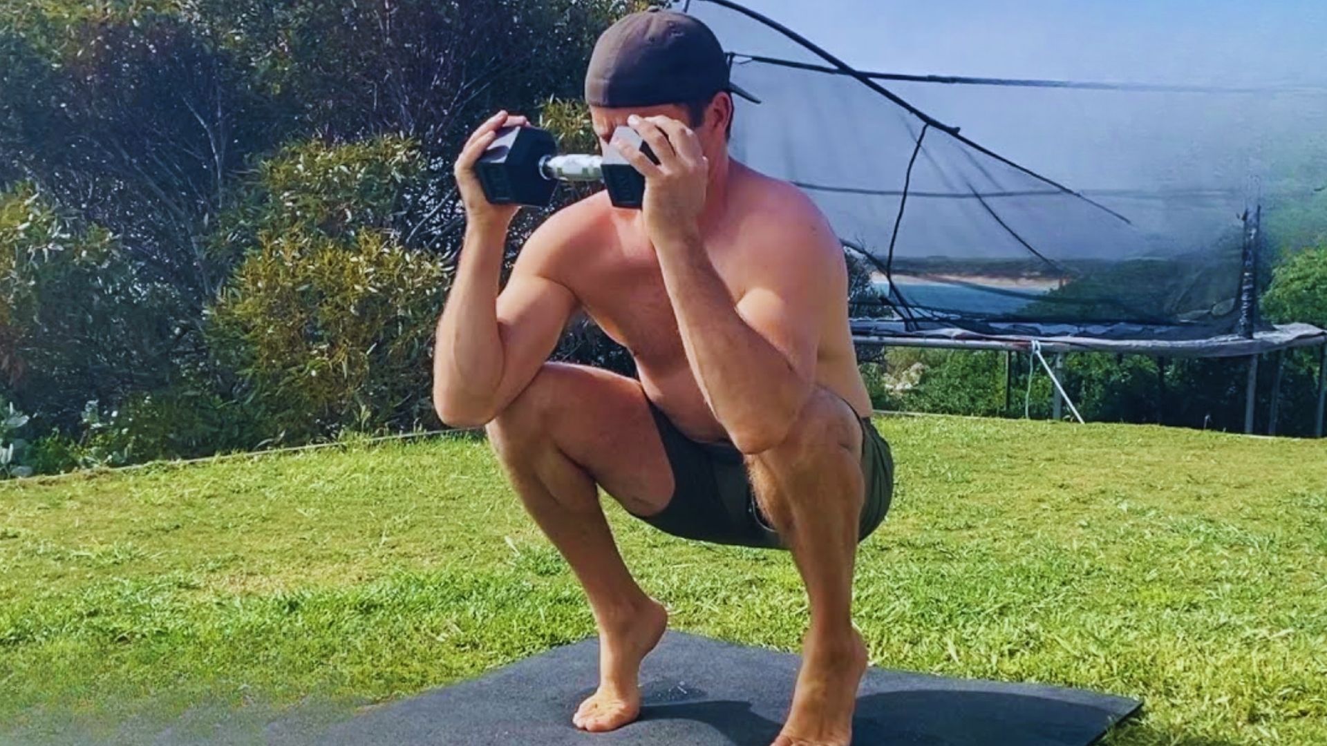 A shirtless man squats on a mat, holding a camera, focused on capturing a photo or video.