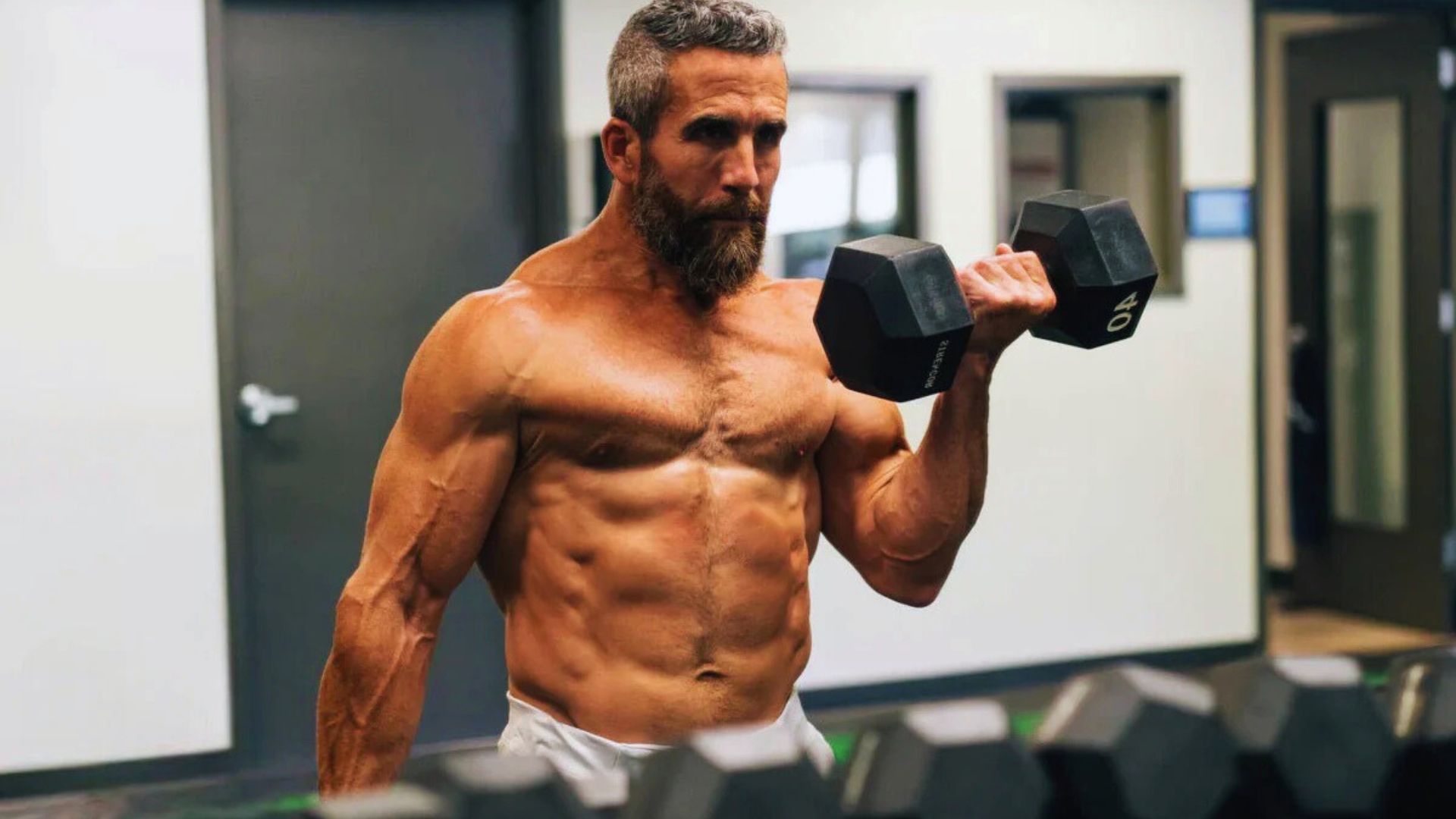 Muscular man in gym lifting a dumbbell with intense focus. The background shows gym equipment and exercise areas, conveying strength and determination.