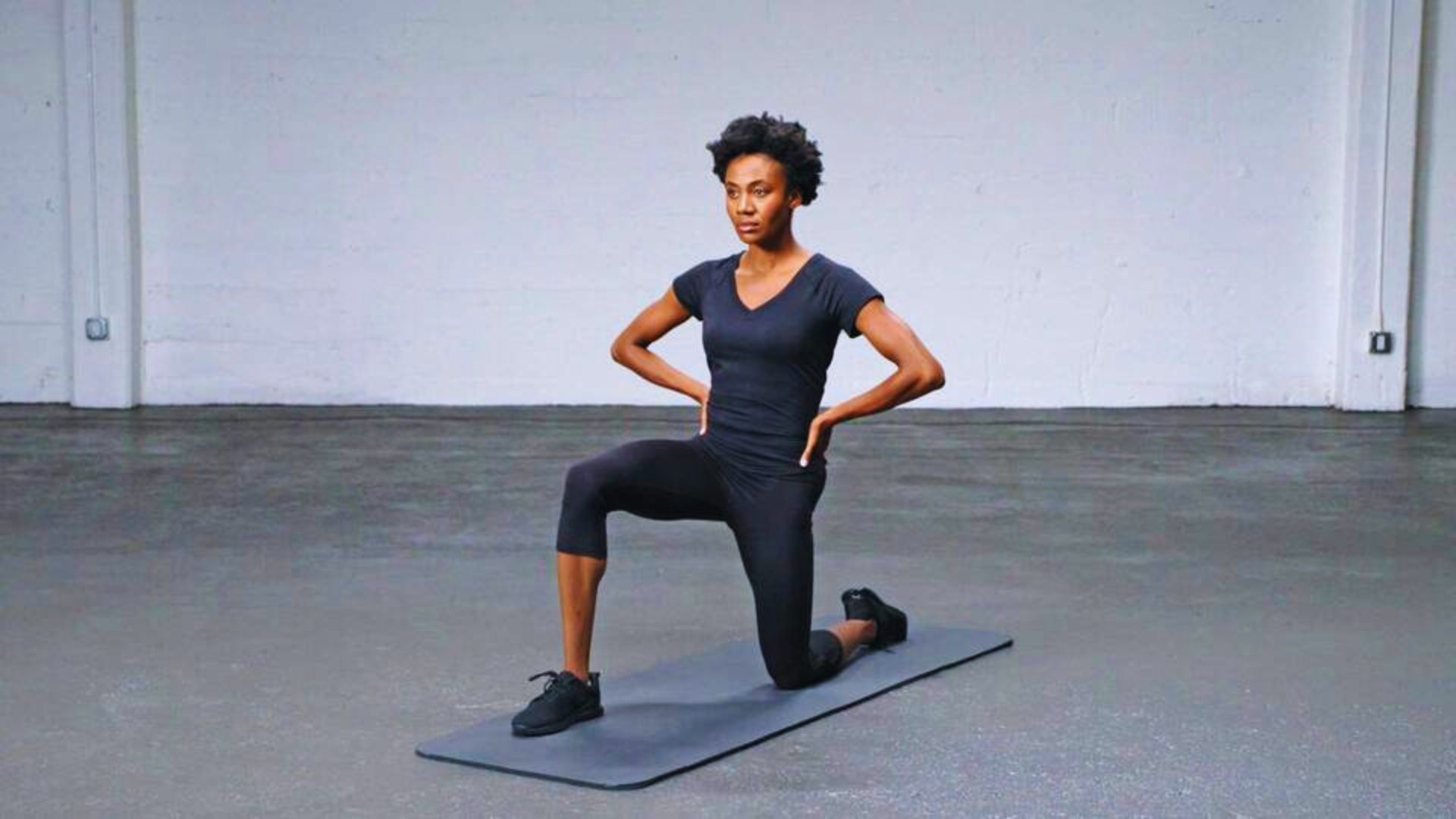 A person in workout attire performs a kneeling lunge on a yoga mat in a spacious room. They look focused, demonstrating balance and strength.