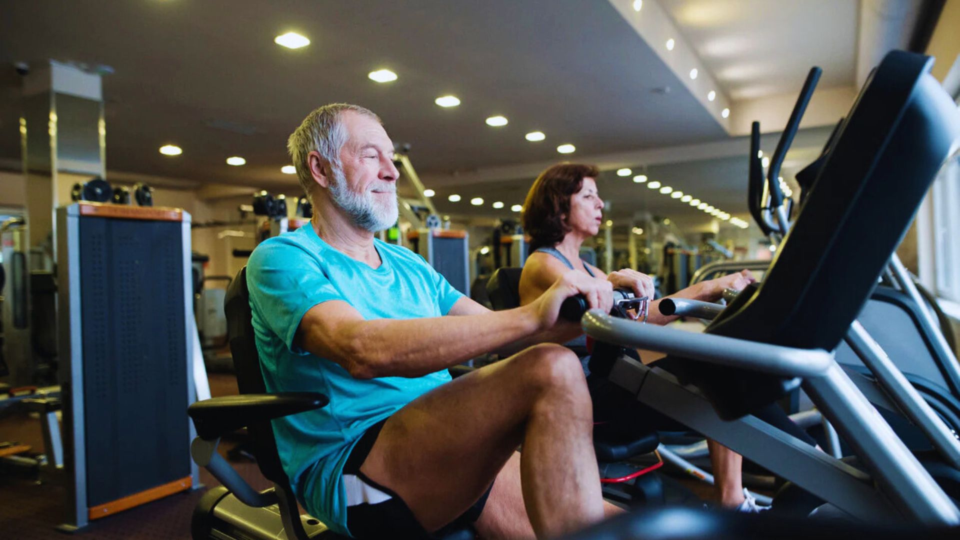 Older man and woman exercising on stationary bikes in a gym. The man wears a blue shirt and shorts, while the woman sports workout attire. Both look focused.