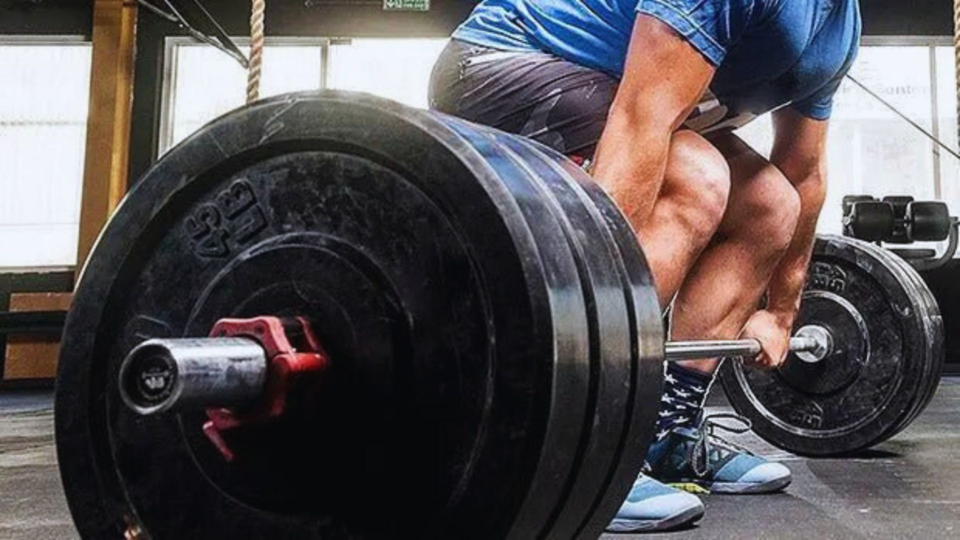A person in a blue shirt and shorts is deadlifting heavy barbells in a gym. The scene conveys strength and focus, highlighting intense effort.