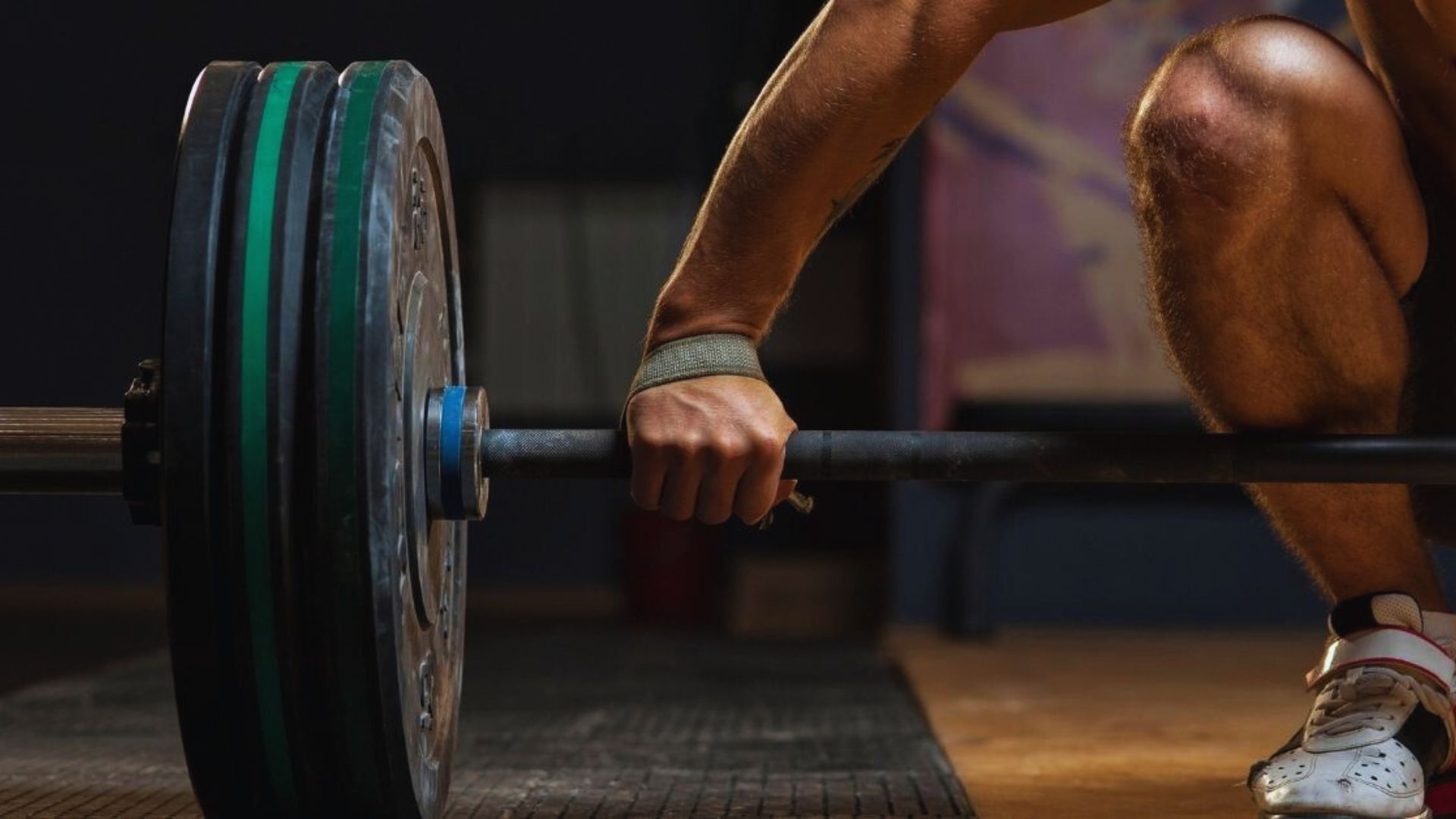 A weightlifter's hand grips a barbell loaded with heavy plates. The scene conveys strength and focus, highlighted by muted gym lighting and intense atmosphere.