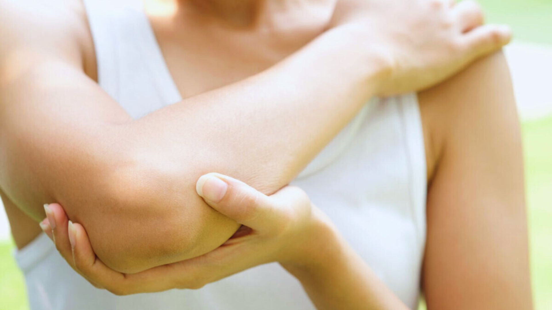 A person wearing a white sleeveless top holds their elbow with the opposite hand, suggesting discomfort or pain. The background is softly blurred and green.