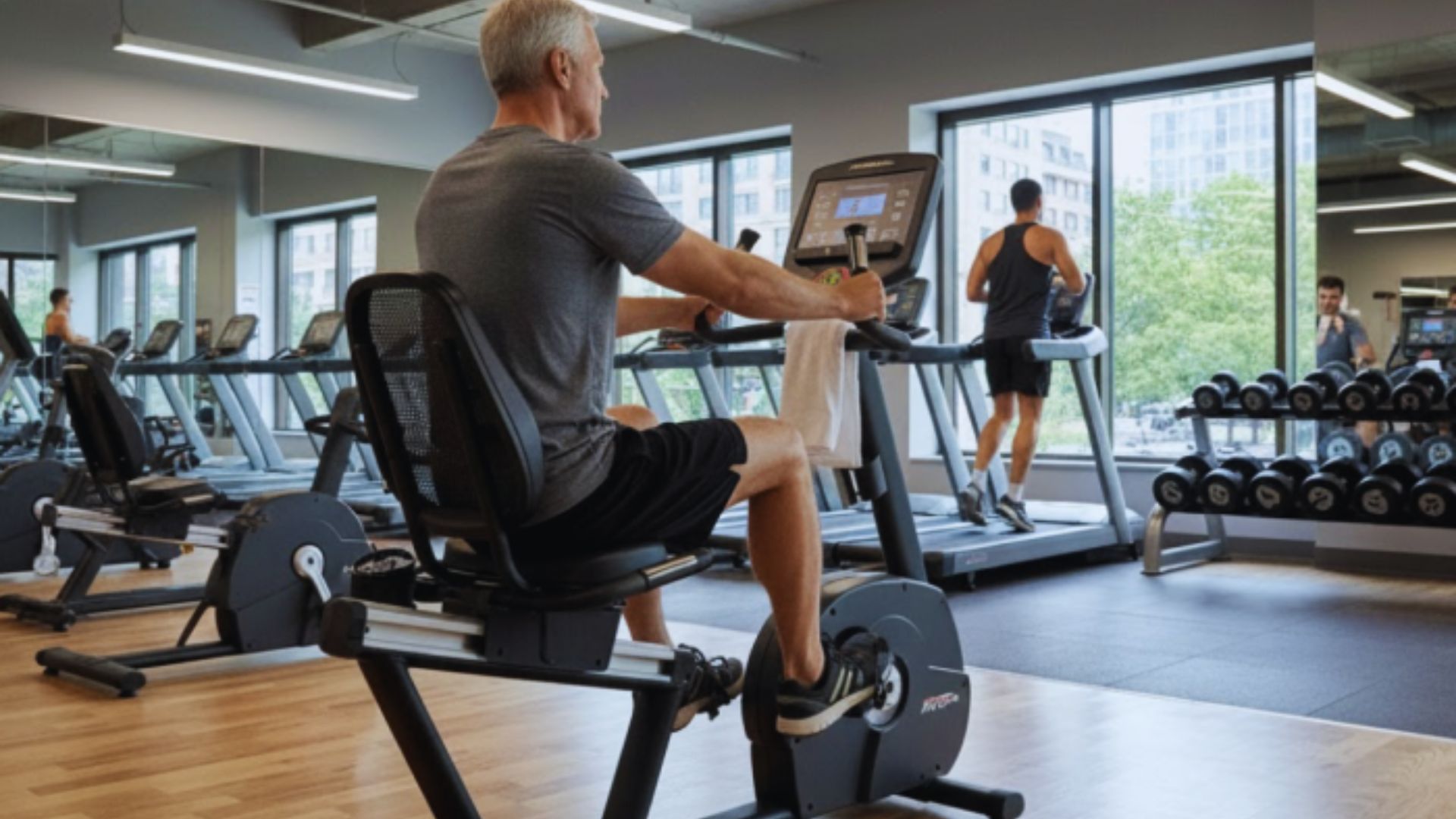 A man with gray hair exercises on a stationary bike in a gym with large windows, while others use treadmills. The atmosphere is focused and active.
