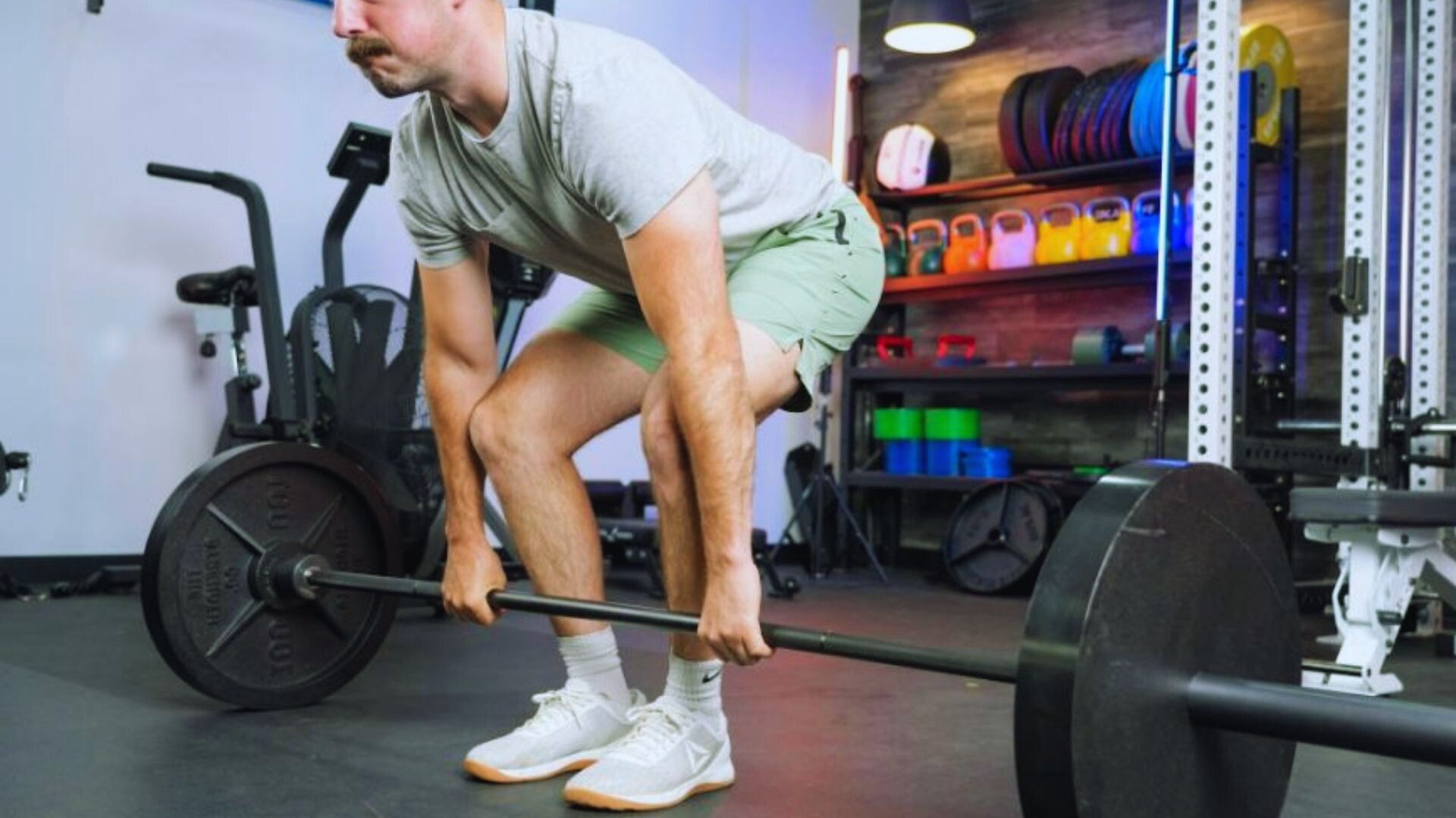 A man in a light gray t-shirt and green shorts prepares to lift a heavy barbell in a gym. Behind him are colorful kettlebells and fitness equipment.