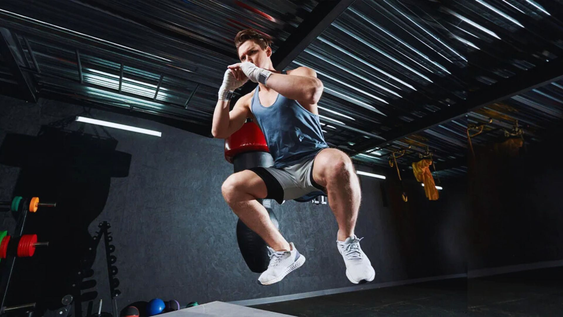 A man in athletic gear jumps toward a punching bag in a dimly lit gym. He wears a blue tank top, gray shorts, and white sneakers, portraying intense focus.