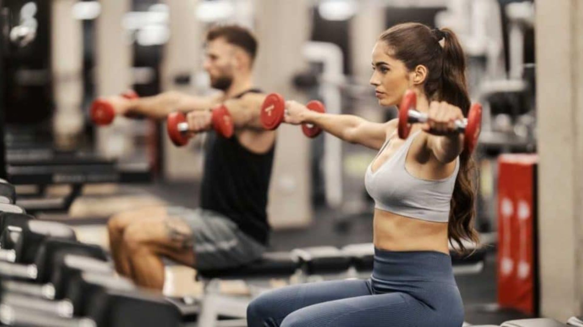 A man and woman perform dumbbell exercises together in a well-equipped gym setting.
