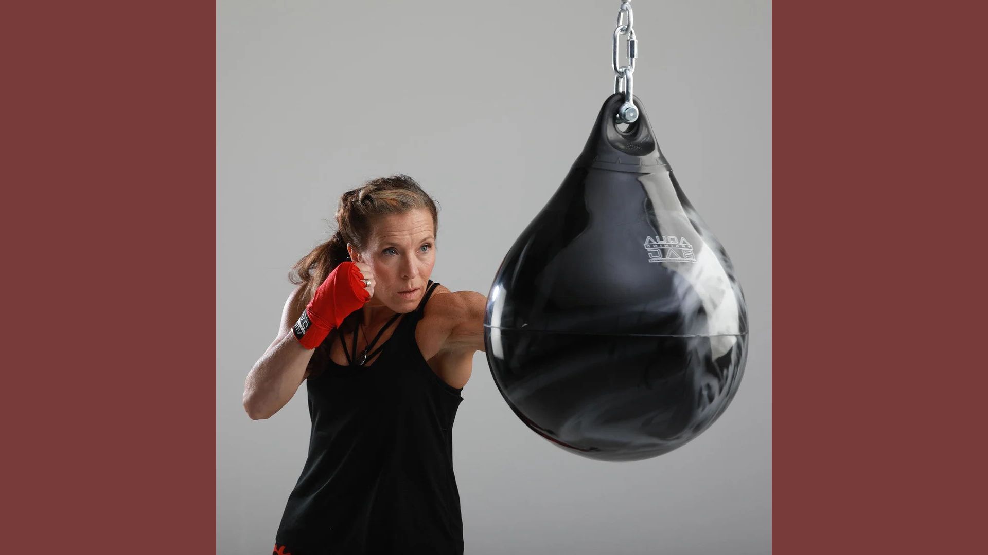 A woman wearing a black shirt and red gloves strikes a punching bag with focused intensity.