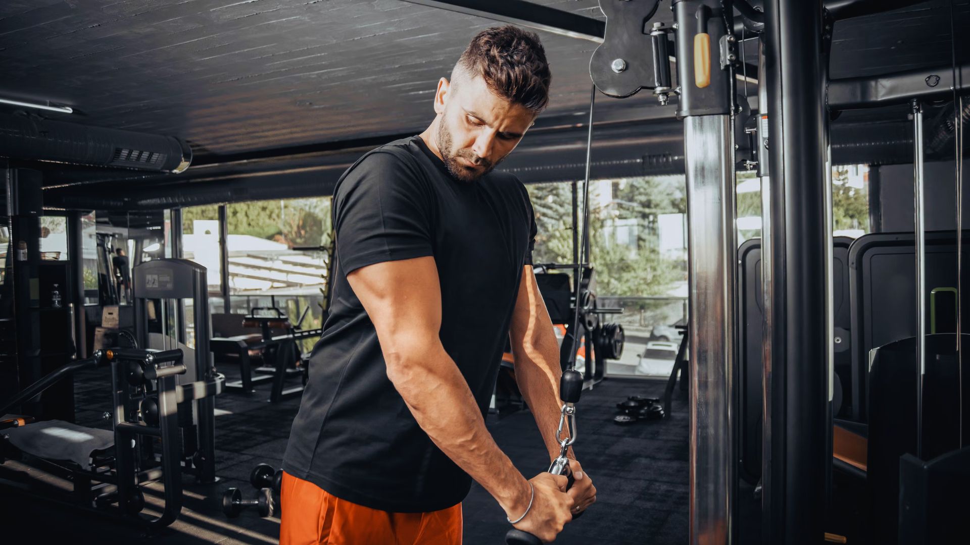 A man in a black T-shirt and orange shorts works out on a cable machine in a gym, focusing on a tricep exercise. The setting is modern and well-lit.
