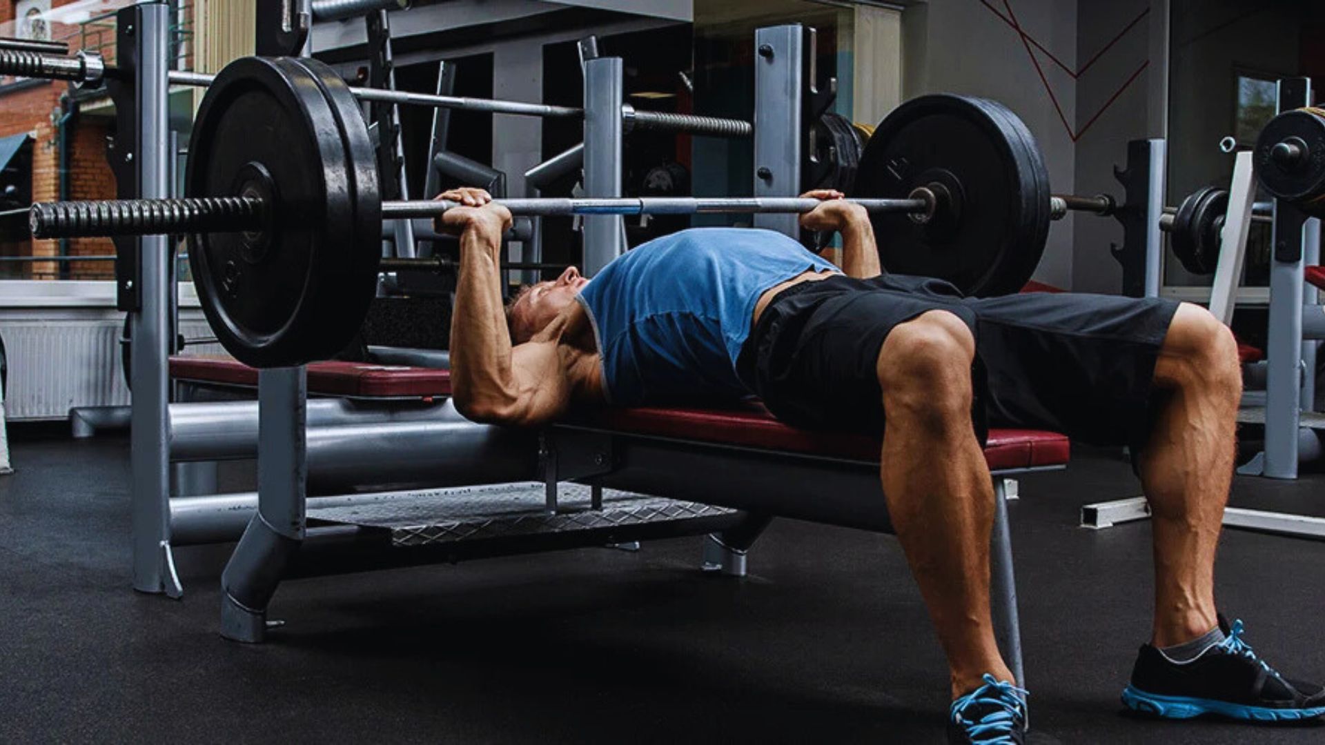 A person in a blue tank top and black shorts is bench pressing a loaded barbell at a gym, showcasing strength and focus in a modern workout environment.