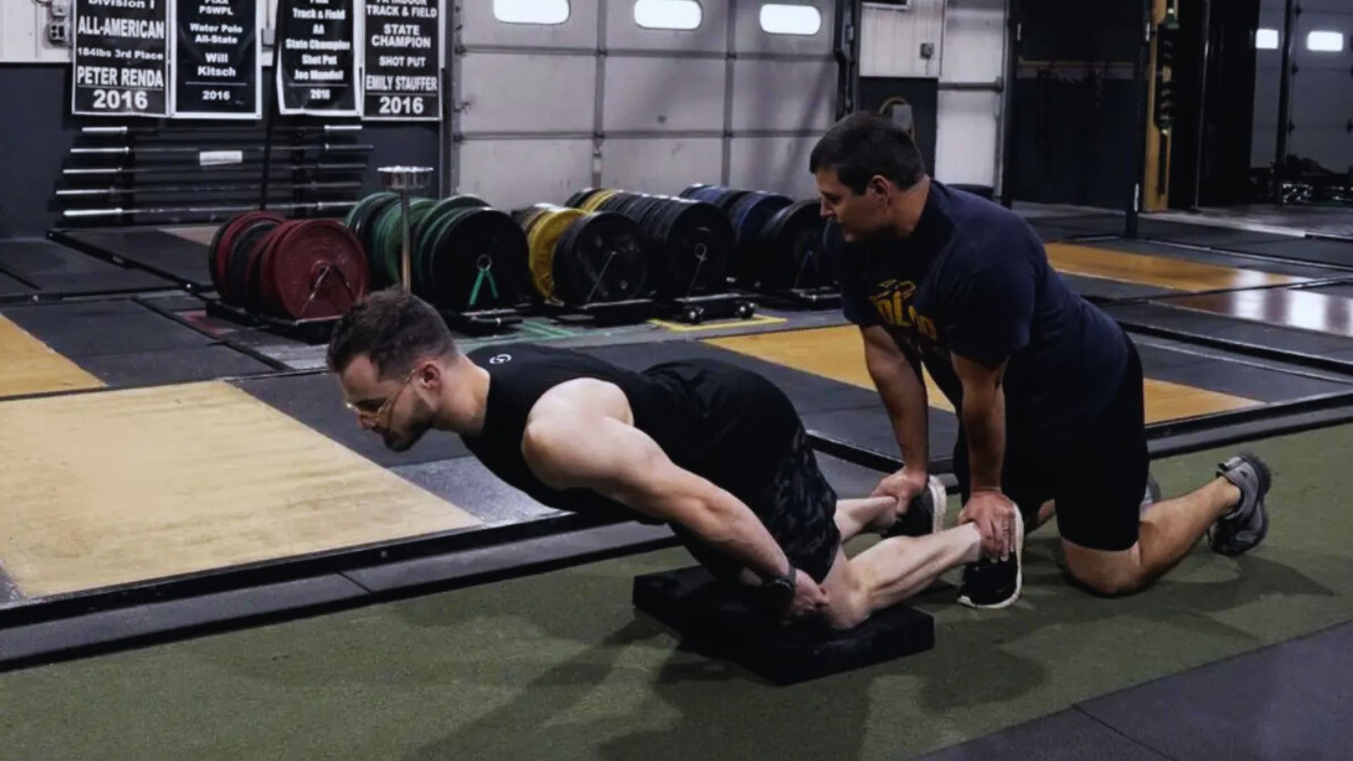 A man performs a Nordic hamstring curl, supported by another man holding his feet. They're in a gym with colorful weight plates on racks behind them.