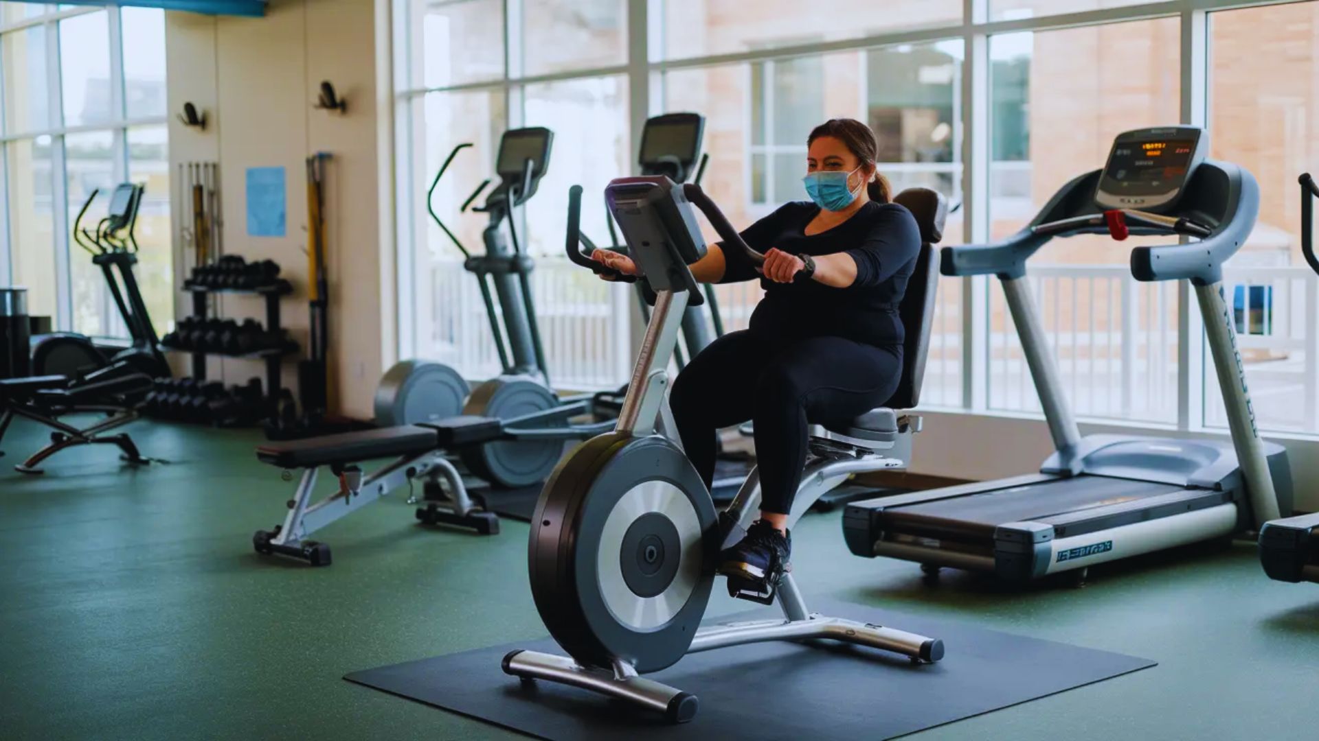 A woman wearing a face mask pedals on an exercise bike in a fitness setting.
