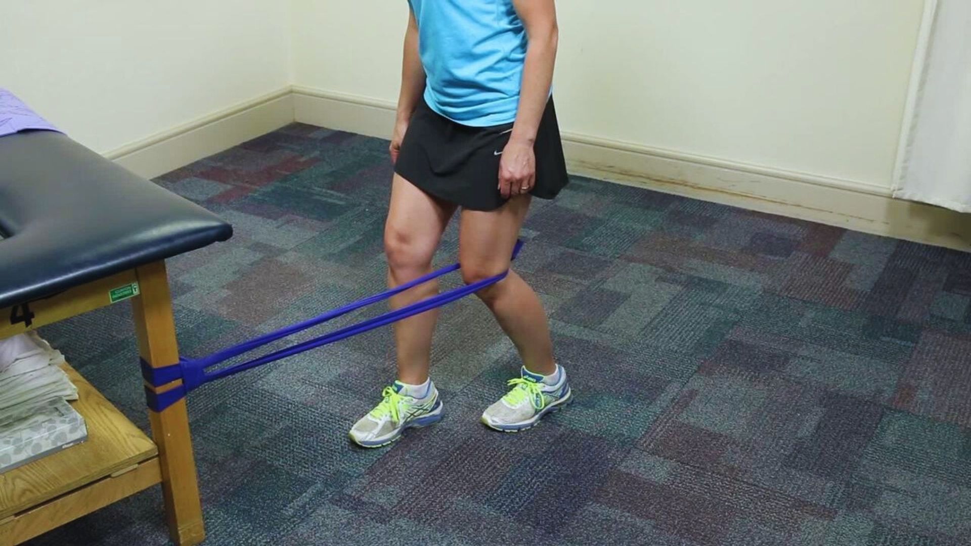 A person wearing a blue shirt and black shorts uses a blue resistance band for exercise, stretching it from a table to their ankle in a room with a carpeted floor.