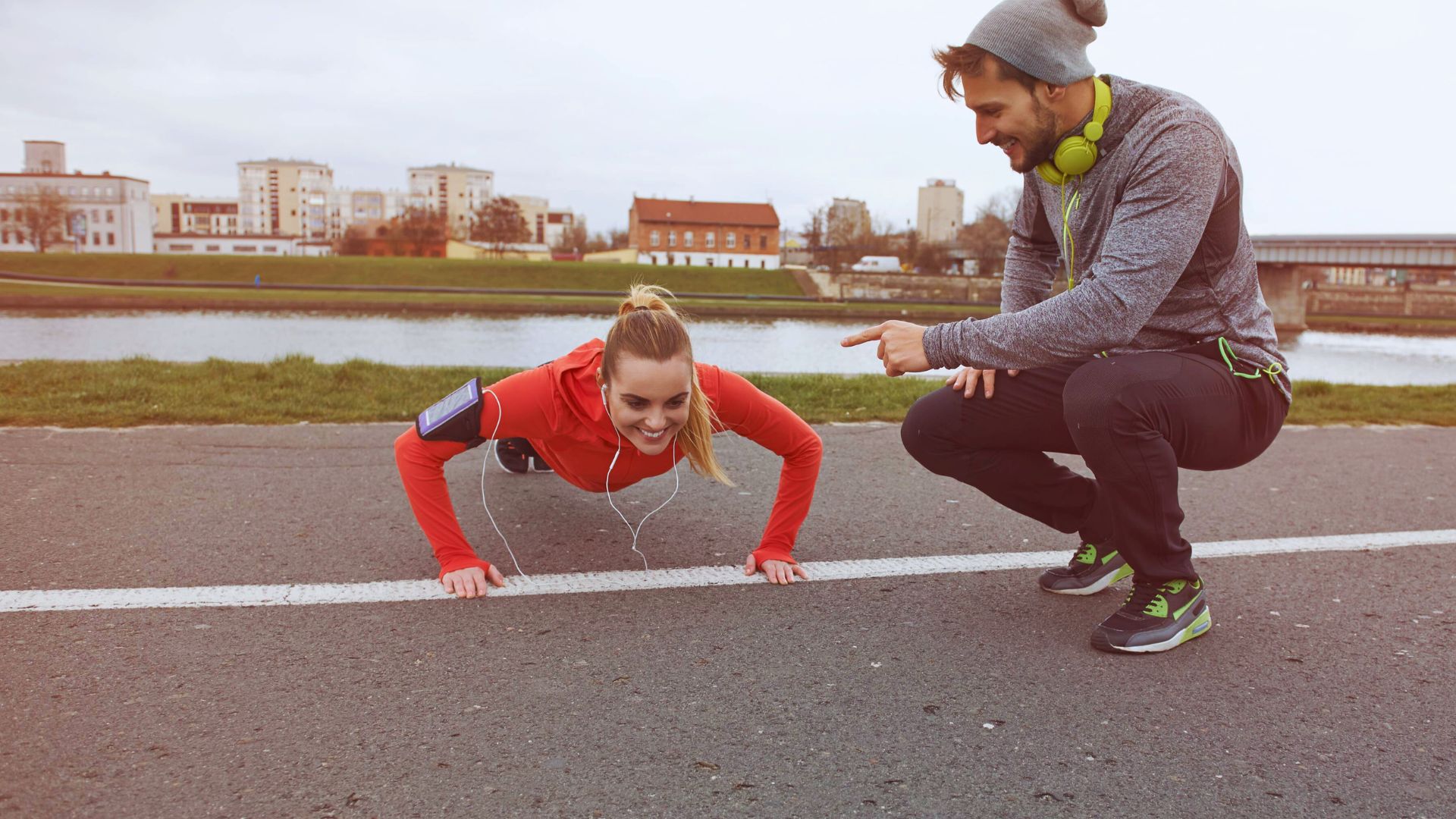 A woman in a red hoodie does push-ups on a riverside path, smiling, while a man crouches nearby, encouraging her. The sky is overcast, and buildings line the horizon.