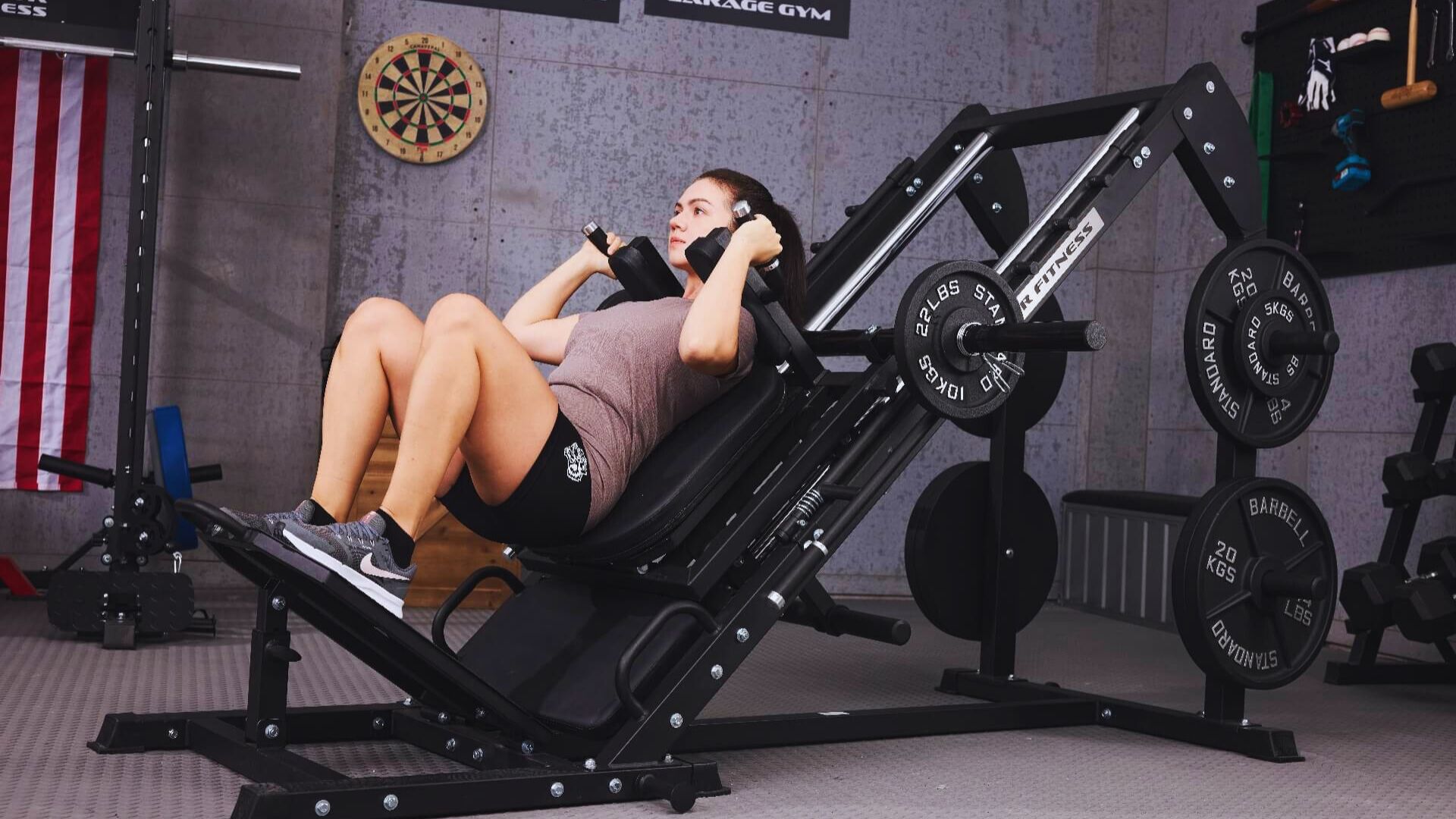 Woman exercising on a leg press machine in a gym, demonstrating strength and focus. A dartboard and American flag are visible in the background.