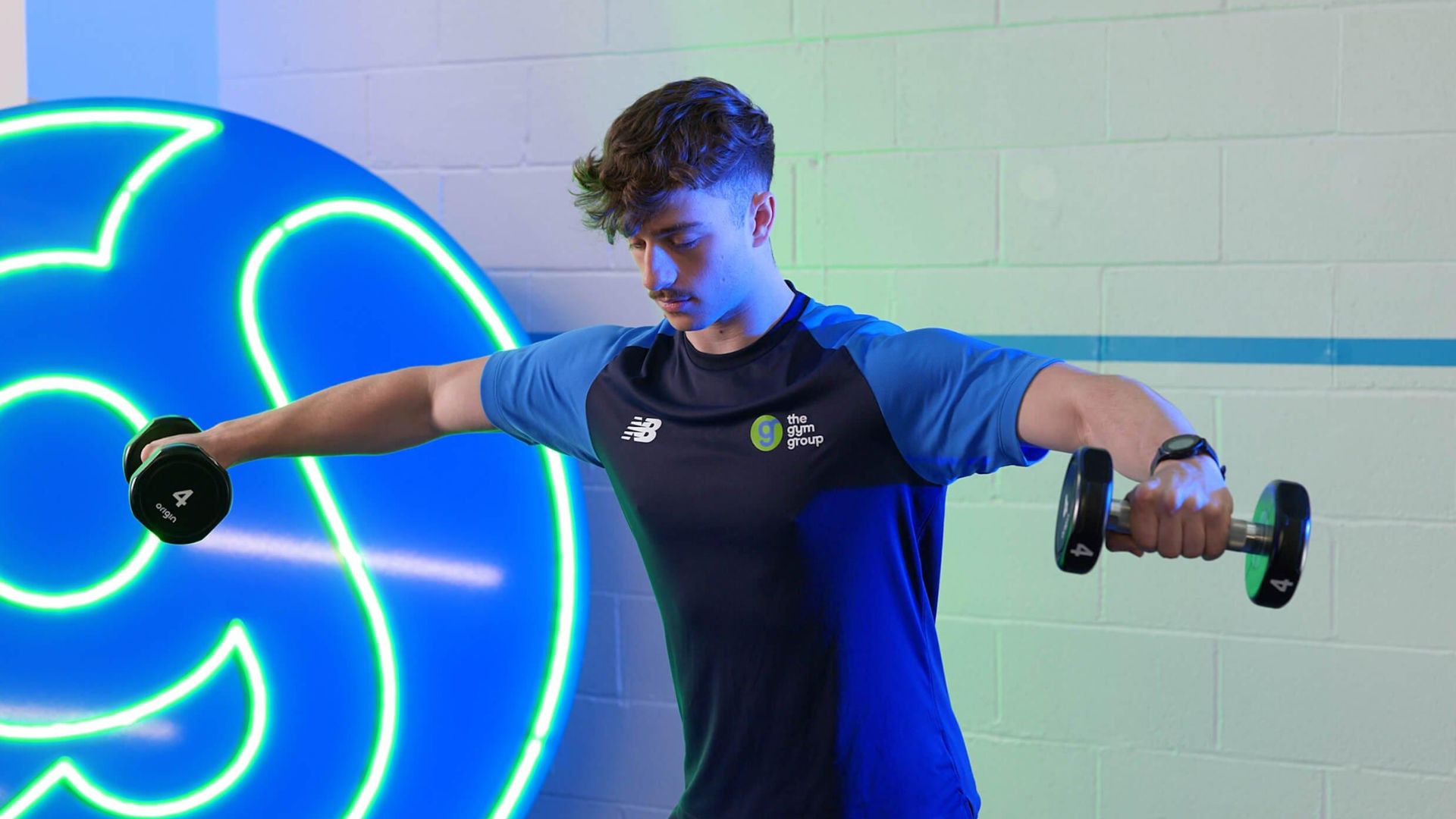 A man performs a workout, lifting two dumbbells in a gym setting.