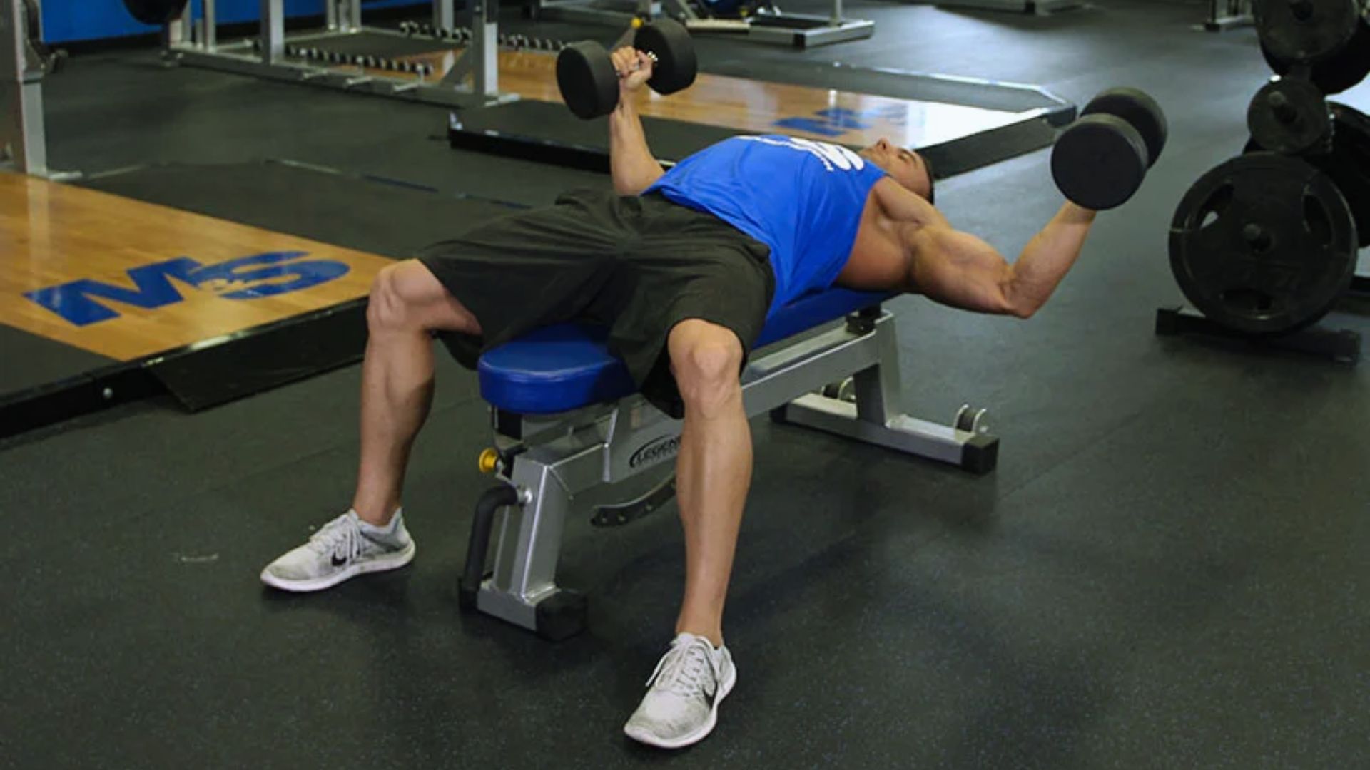 A man performs a bench press exercise using dumbbells in a gym setting.
