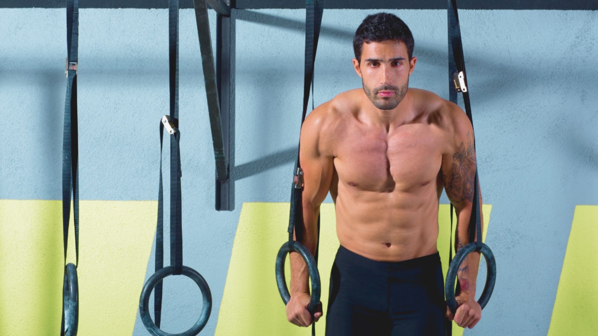 A shirtless man holds a ring while standing on a cross trainer in a gym setting.