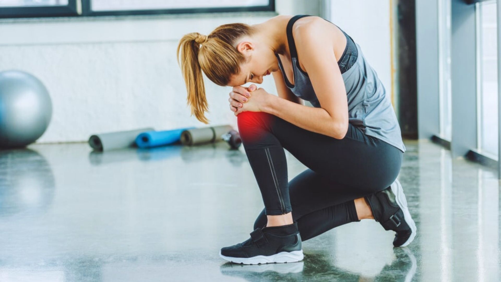 A woman kneels indoors with a concerned expression, clutching her knee highlighted in red. Yoga mats and exercise ball are in the background.