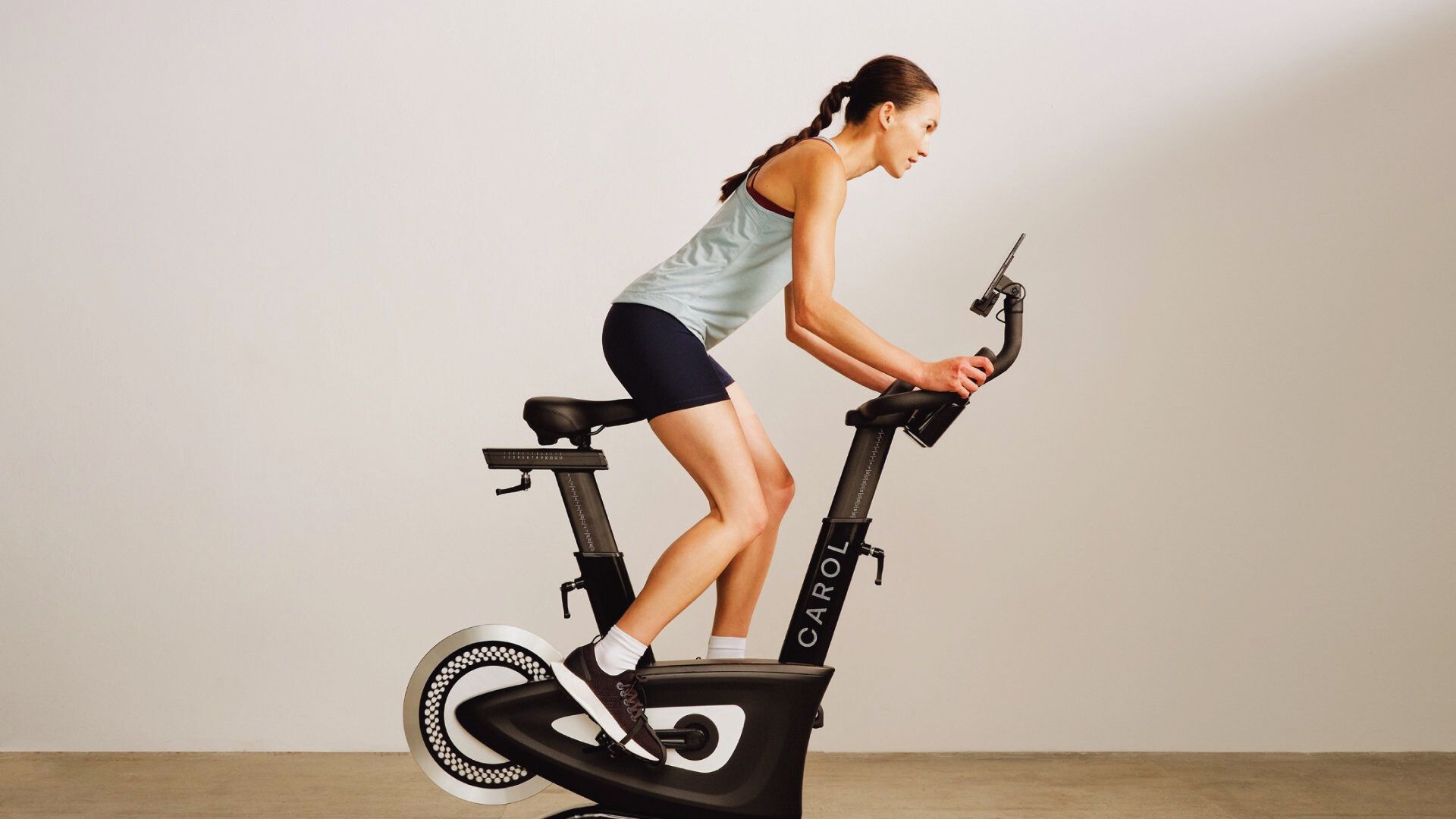 A woman in athletic wear rides an exercise bike indoors. The room is softly lit, and the woman appears focused and determined as she pedals.