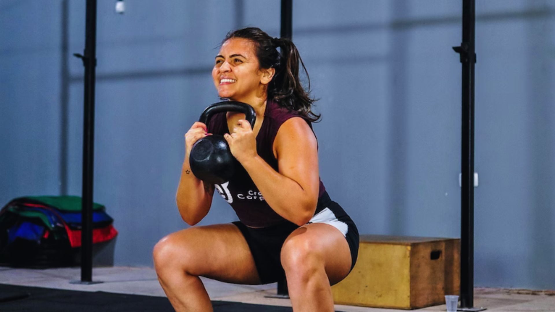 A woman in workout attire performs a kettlebell squat in a gym. She appears focused and determined. Equipment and a wooden box are in the background.