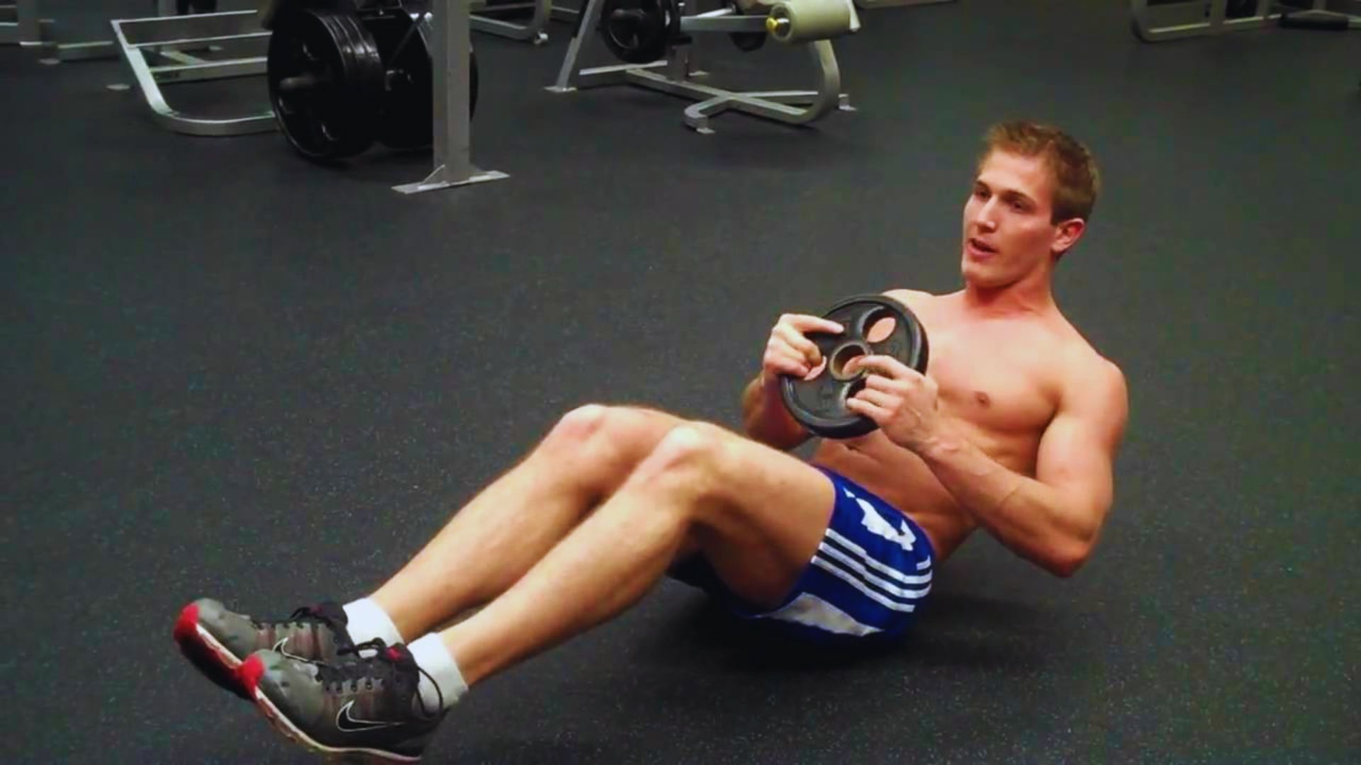 A shirtless man performs a sit-up on a gym floor, focusing on his exercise routine.
