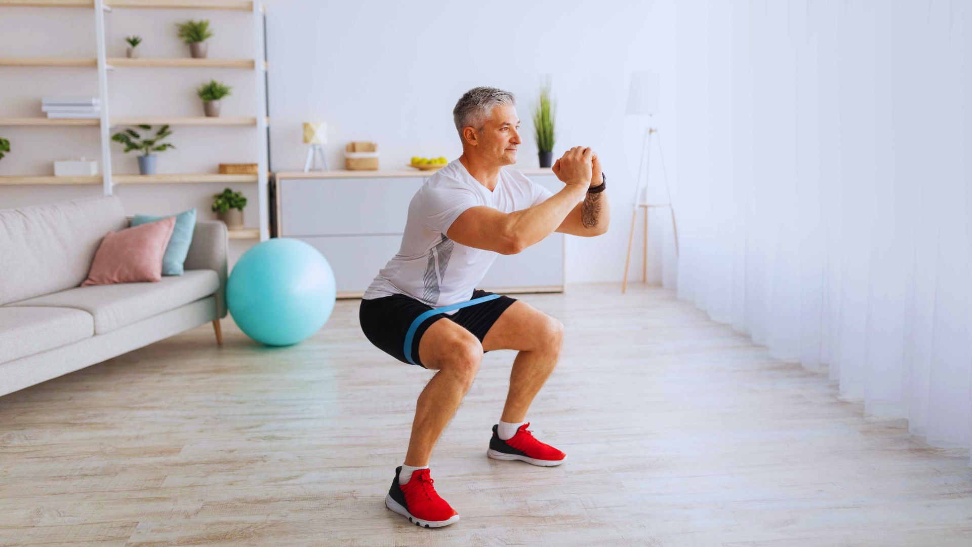 A man exercises in a bright living room, doing squats with a resistance band. He wears a white shirt and red sneakers. A yoga ball and sofa are visible.