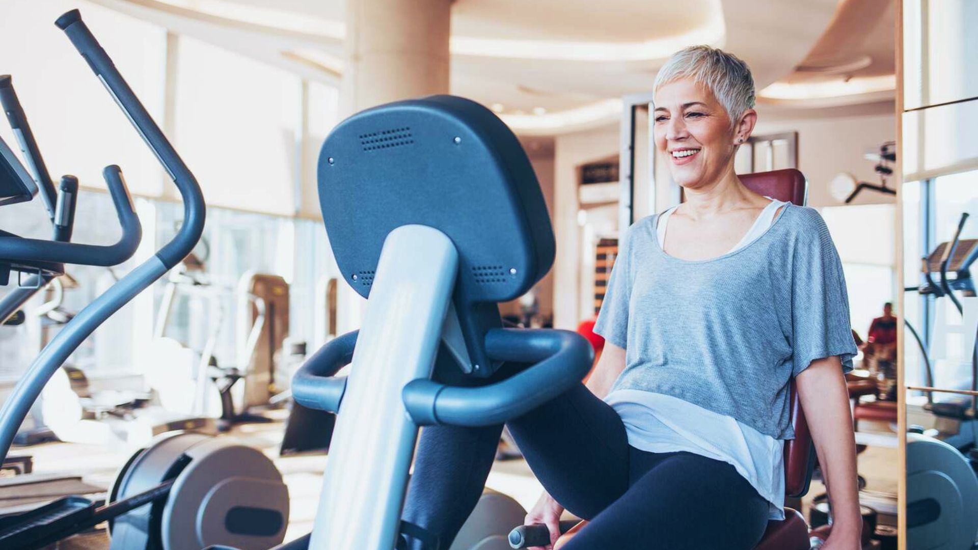 A woman with short hair smiles while exercising on a stationary bike in a bright, spacious gym. The atmosphere is energetic and motivating.