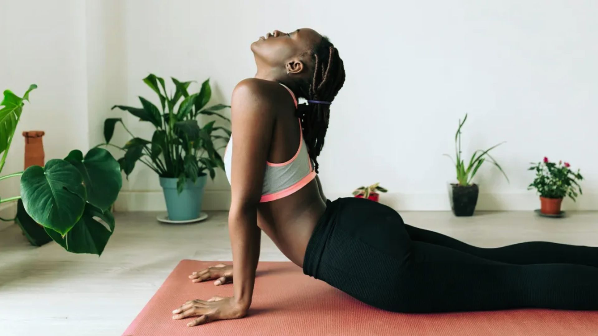 A person practices yoga, performing a cobra pose on a mat. They are in a serene room with green potted plants, wearing a white sports top and black leggings.