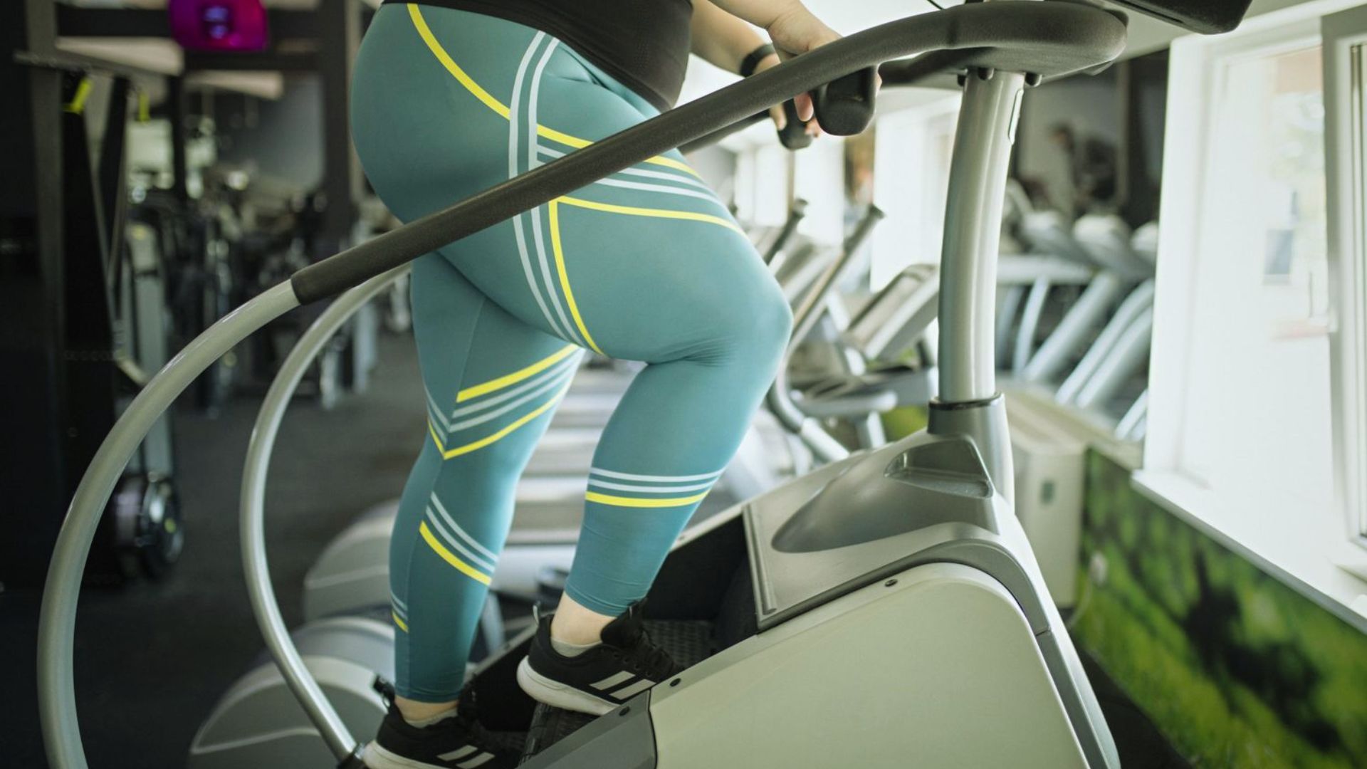 A person wearing blue leggings with yellow stripes exercises on a stair stepper in a bright gym. The focus is on their legs and the machine.