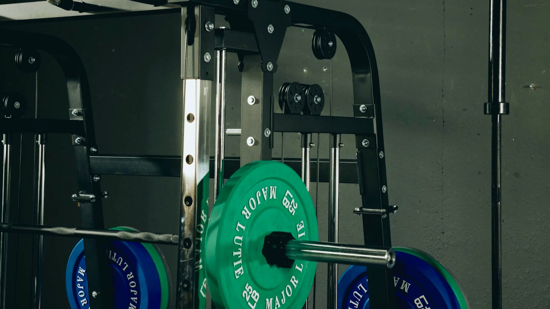 A gym setting with a Smith machine holding a barbell loaded with green and blue weight plates. The ambiance is industrial and serious.