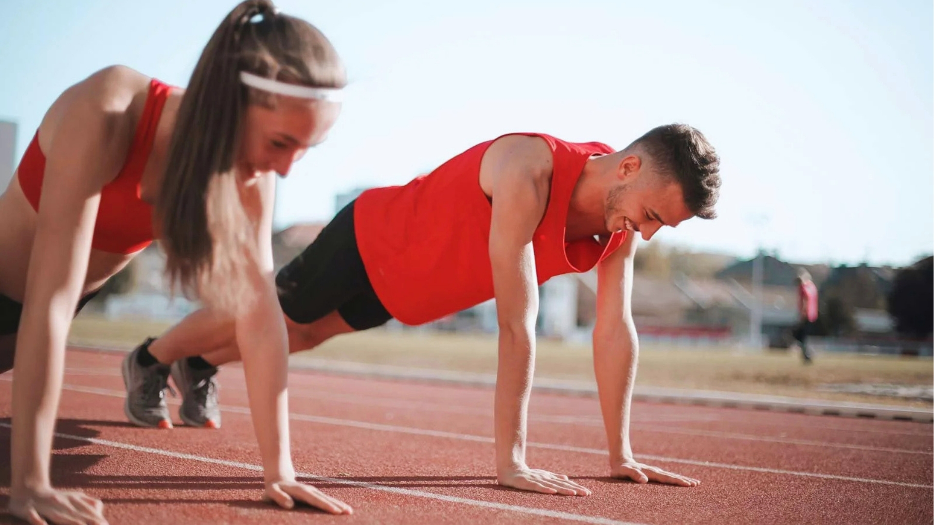 Two people in matching red athletic wear perform push-ups on a sunlit outdoor track, conveying focus and determination in a fitness setting.