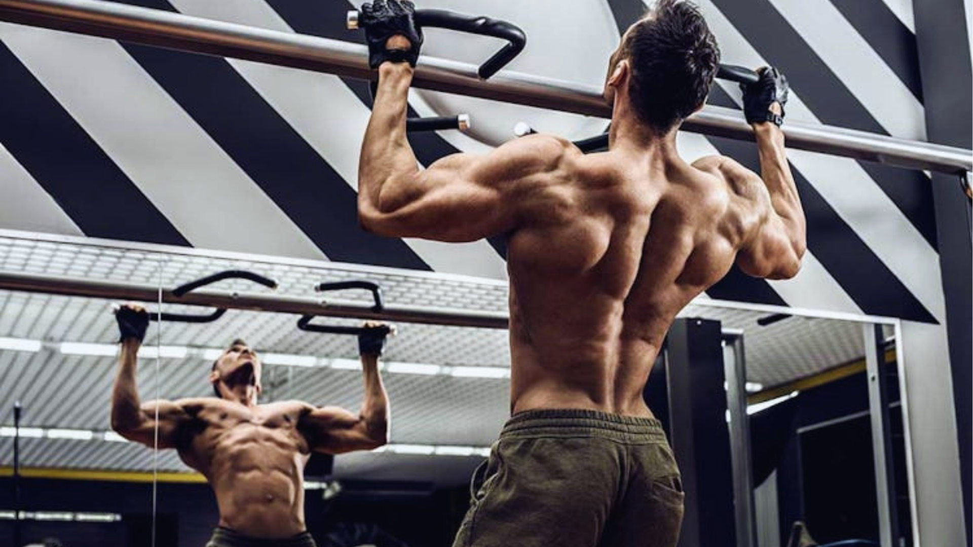 A muscular man performs a pull-up in a gym, facing a mirror. He wears gloves and workout pants. The setting features modern, geometric wall designs.