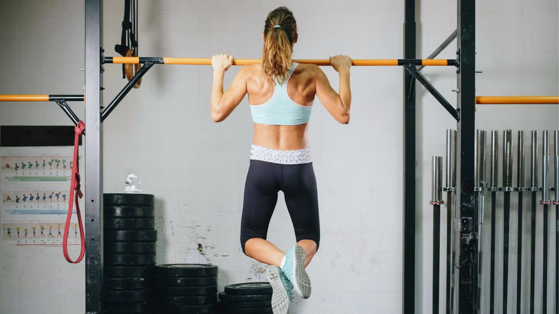 A woman in athletic gear performs a pull-up in a gym. She wears a light blue top and black leggings. Plates and bars are visible; the tone is focused.