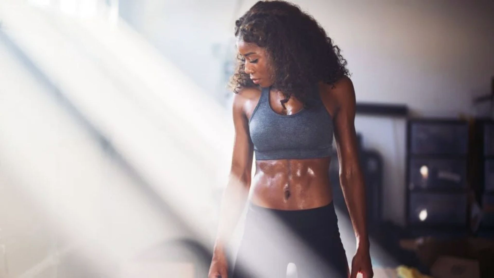 A woman in athletic wear stands in a gym, looking down. Sunlight beams highlight her muscular physique, conveying strength and focus.