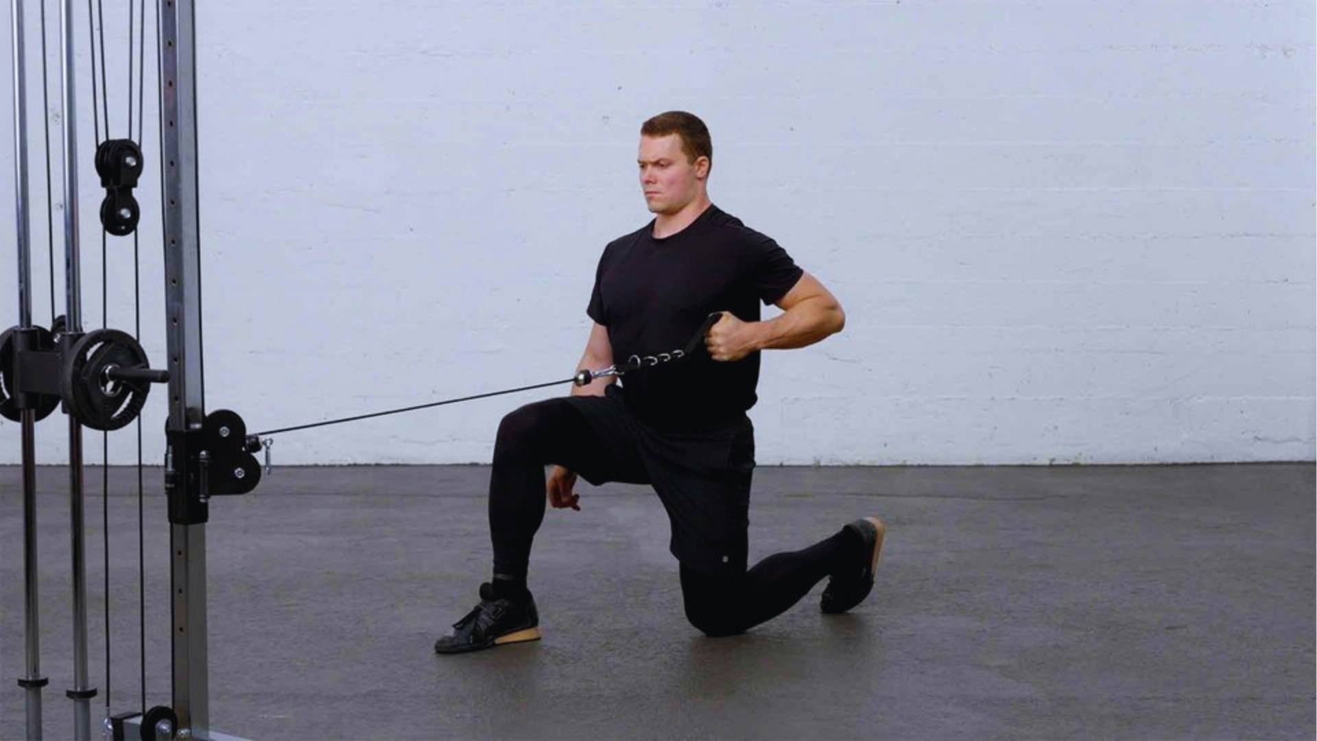 A person kneels on one knee while pulling on a cable machine at a gym, dressed in black athletic wear. The focused expression suggests concentration.