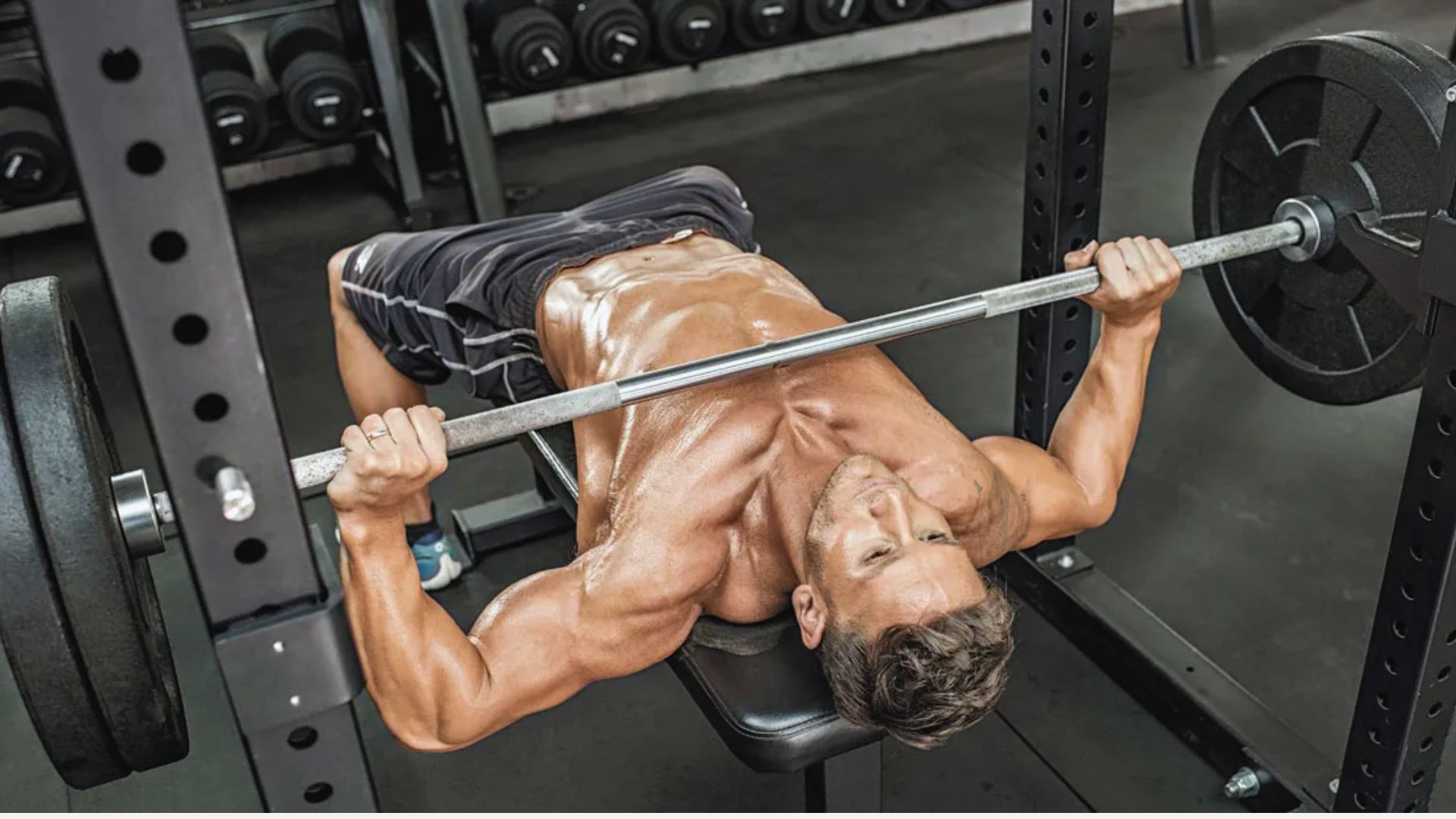 Man bench pressing in a gym, lifting a barbell with heavy weights. Sweat glistens on his muscular physique, conveying strength and determination.