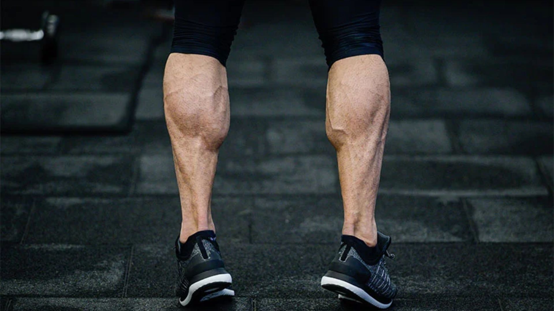 Rear view of muscular calves wearing black sneakers and shorts on a dark gym floor. The scene conveys strength and focus on leg training.
