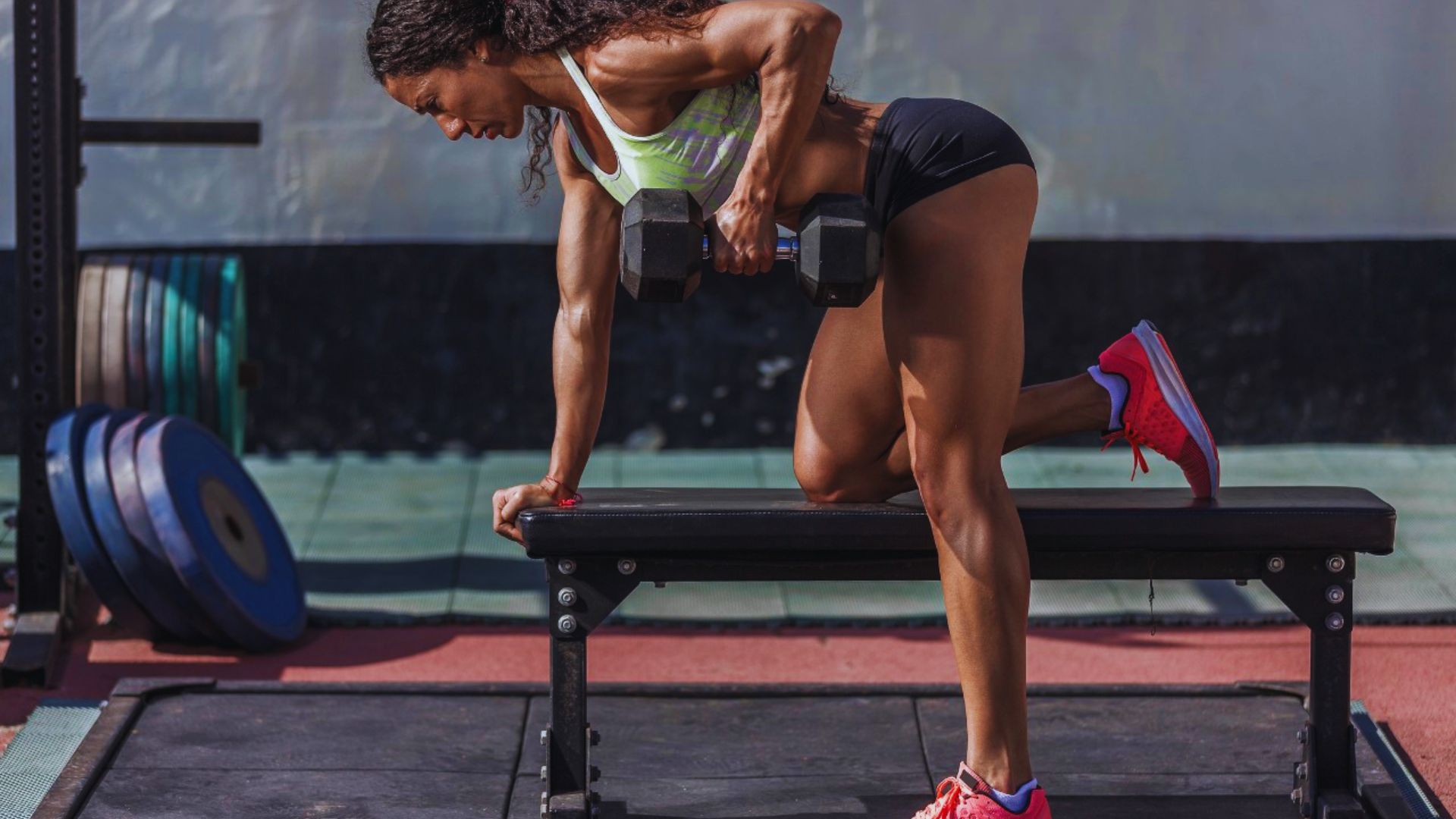 Woman exercising in a gym, performing a dumbbell row while kneeling on a bench. She appears focused and determined, conveying strength and energy.