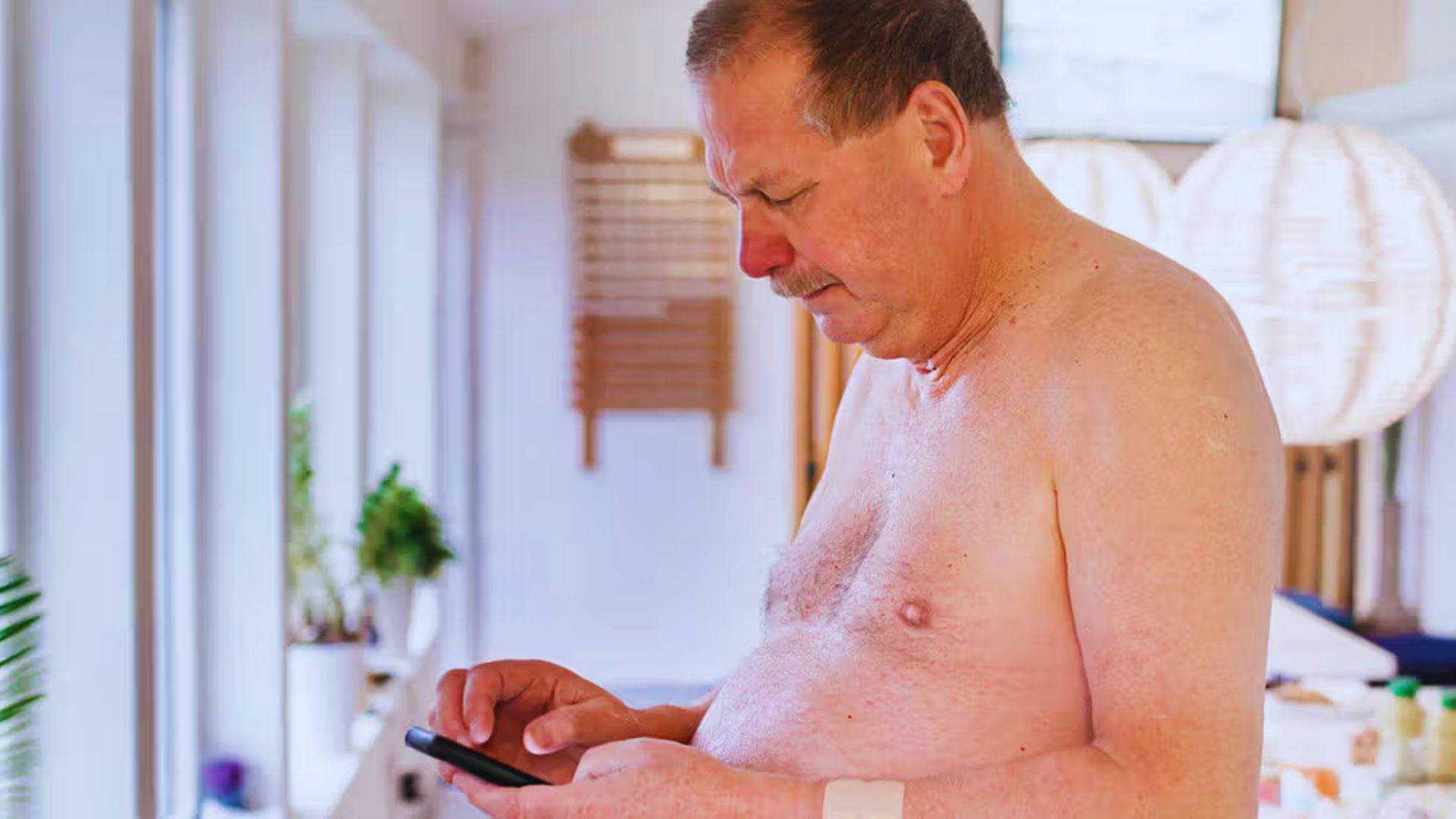 A shirtless man stands indoors, focused on a smartphone. Bright, airy room with plants and round light fixture. Relaxed and casual setting.