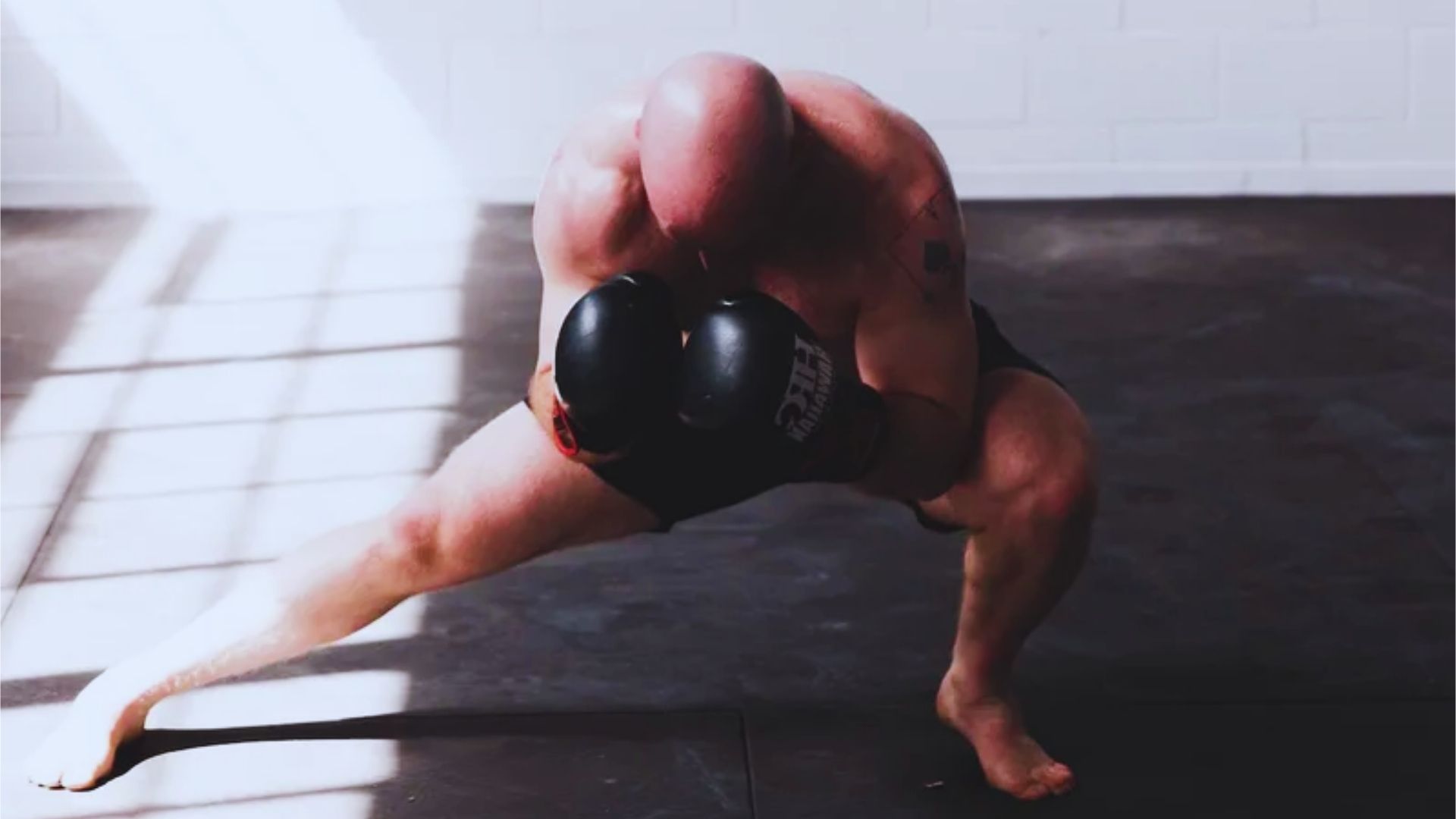 A bald, muscular man practices boxing in a gym with dim lighting. He is crouched, wearing black gloves, conveying strength and focus.