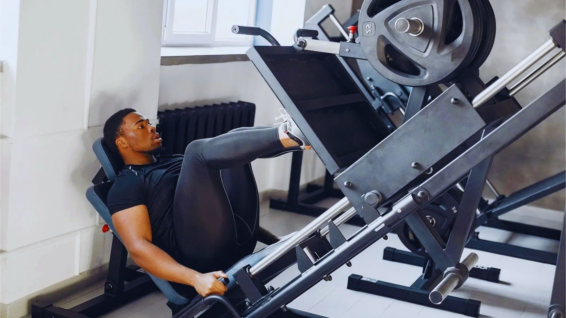 A person exercises on a leg press machine in a gym, focusing intensely. They wear athletic gear, and the setting is bright and modern, conveying determination.
