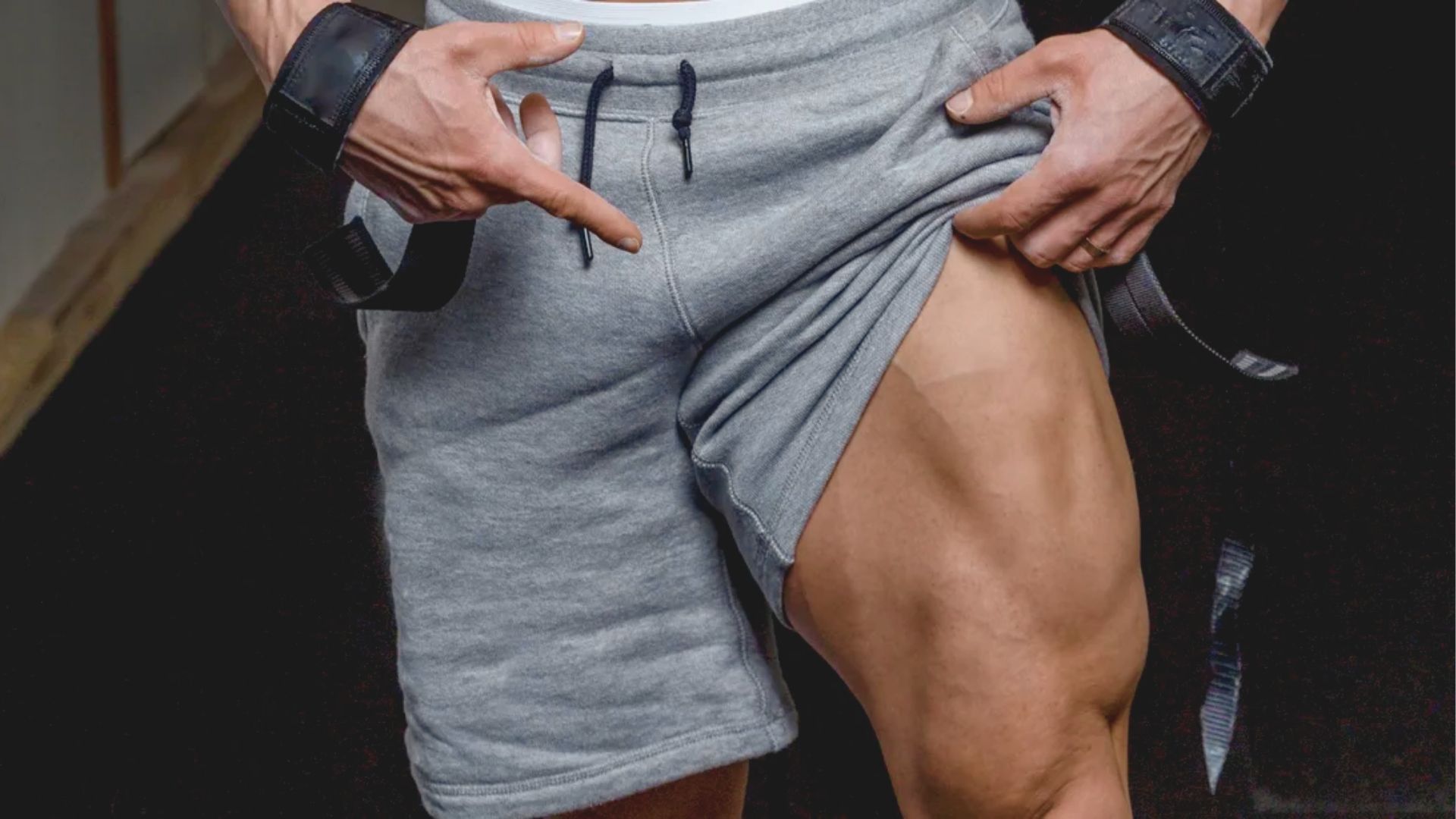 A person in gray shorts points to their muscular thigh, showing prominent muscle definition. The setting appears to be a gym, suggesting strength and fitness.