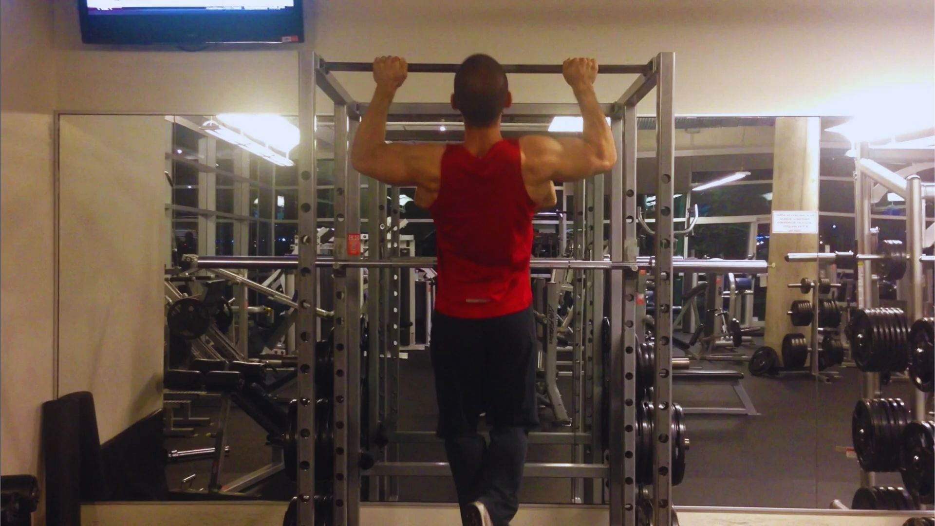 A person in a red tank top performs a pull-up in a gym. The room is equipped with various weightlifting equipment, and a TV is mounted on the wall.